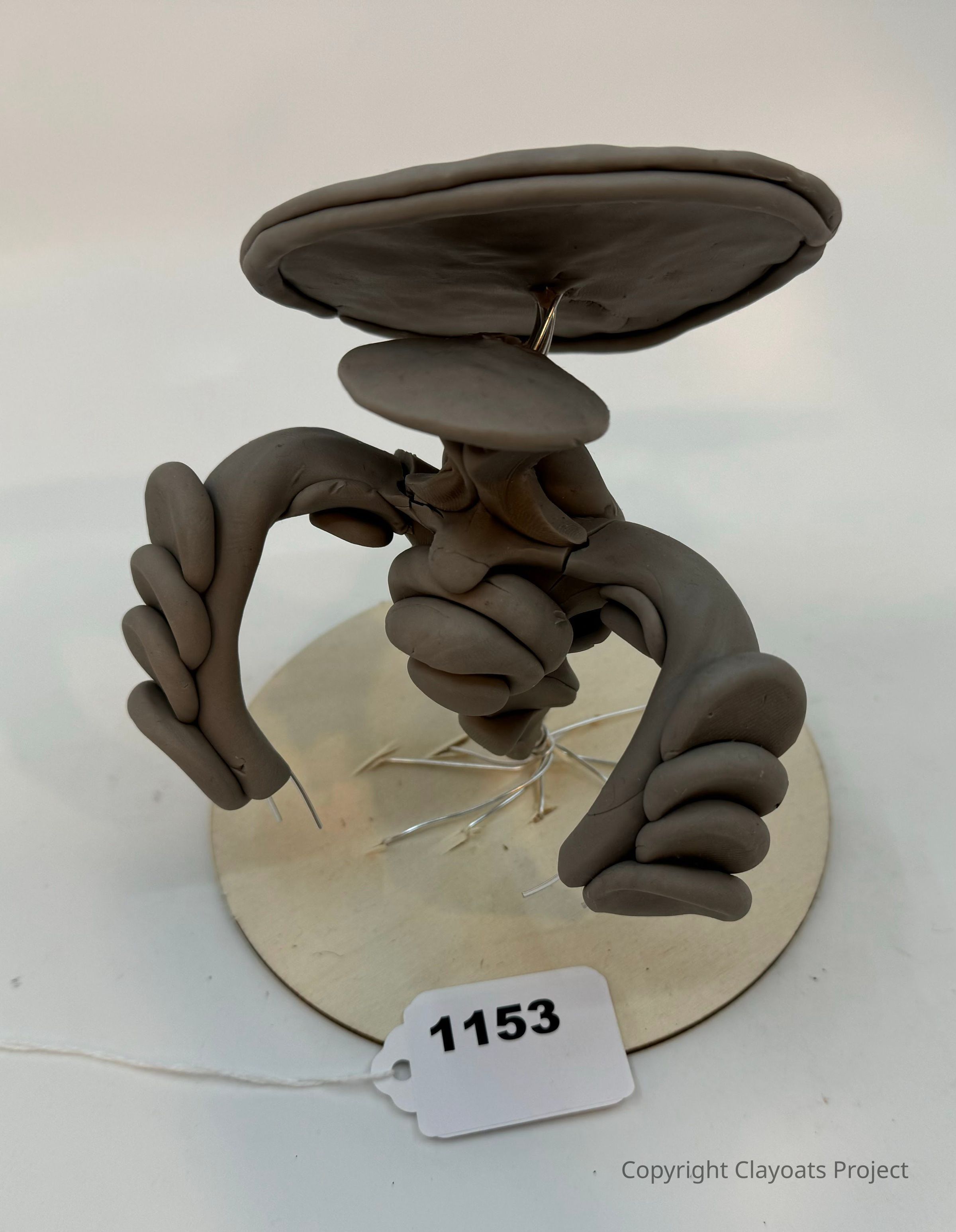 The image showcases a creatively sculpted abstract form made from clay, resting on a circular base with visible wire supports. The sculpture features limb-like structures and a large, flat, disc-shaped element on top, resembling an unconventional figure or creature in a dynamic pose. The surface texture and neutral color of the clay contribute to its minimalist aesthetic.