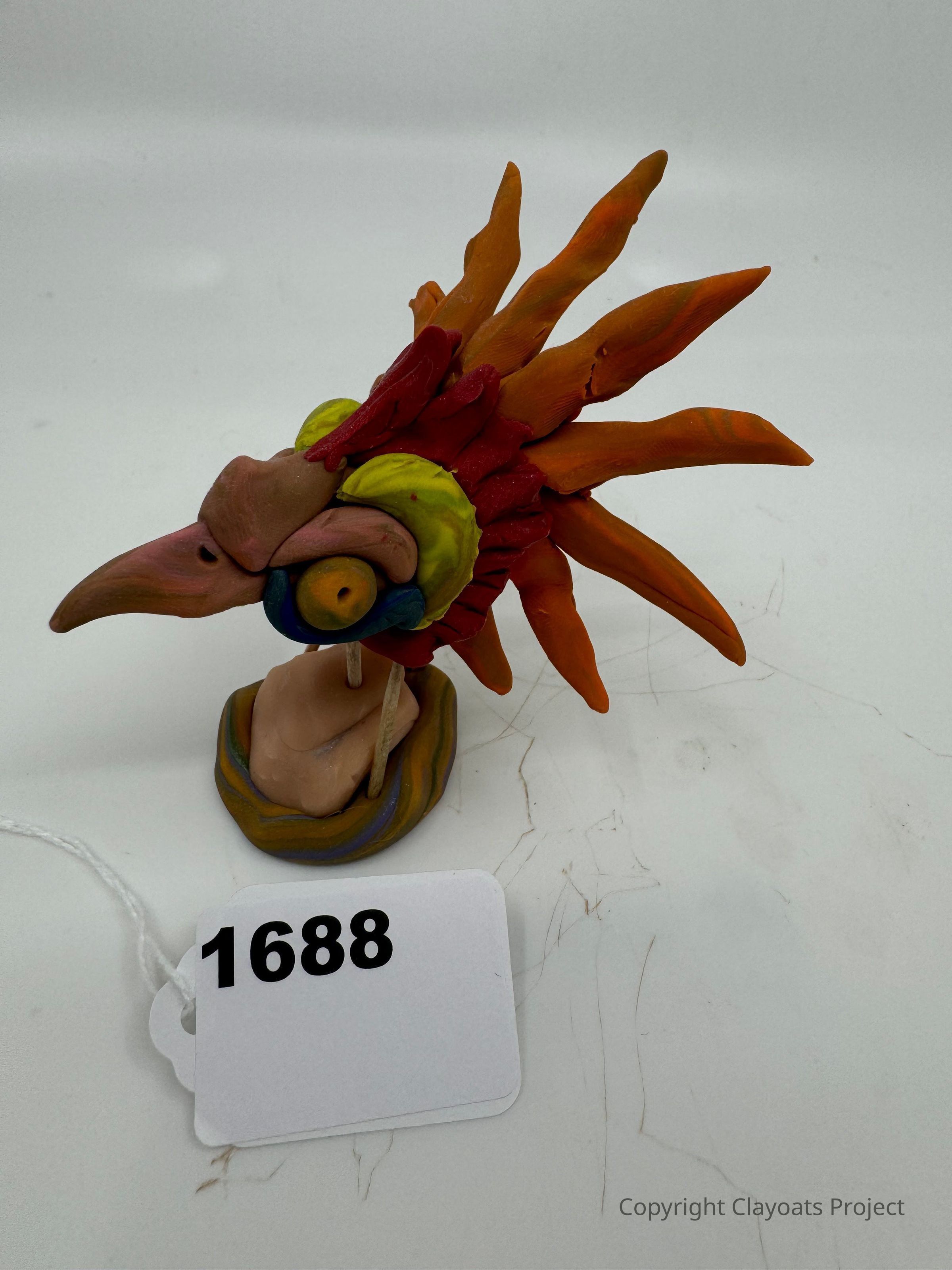 The image shows a colorful and abstract bird-like sculpture made of clay, featuring a prominent beak and vibrant head feathers in shades of orange, red, yellow, and green. The sculpture sits on a small base, set against a plain white background, capturing the intricate details and artistic craftsmanship of the piece. The feathers appear to radiate outward, giving the sculpture an energetic and dynamic look, while the base provides stability, balancing