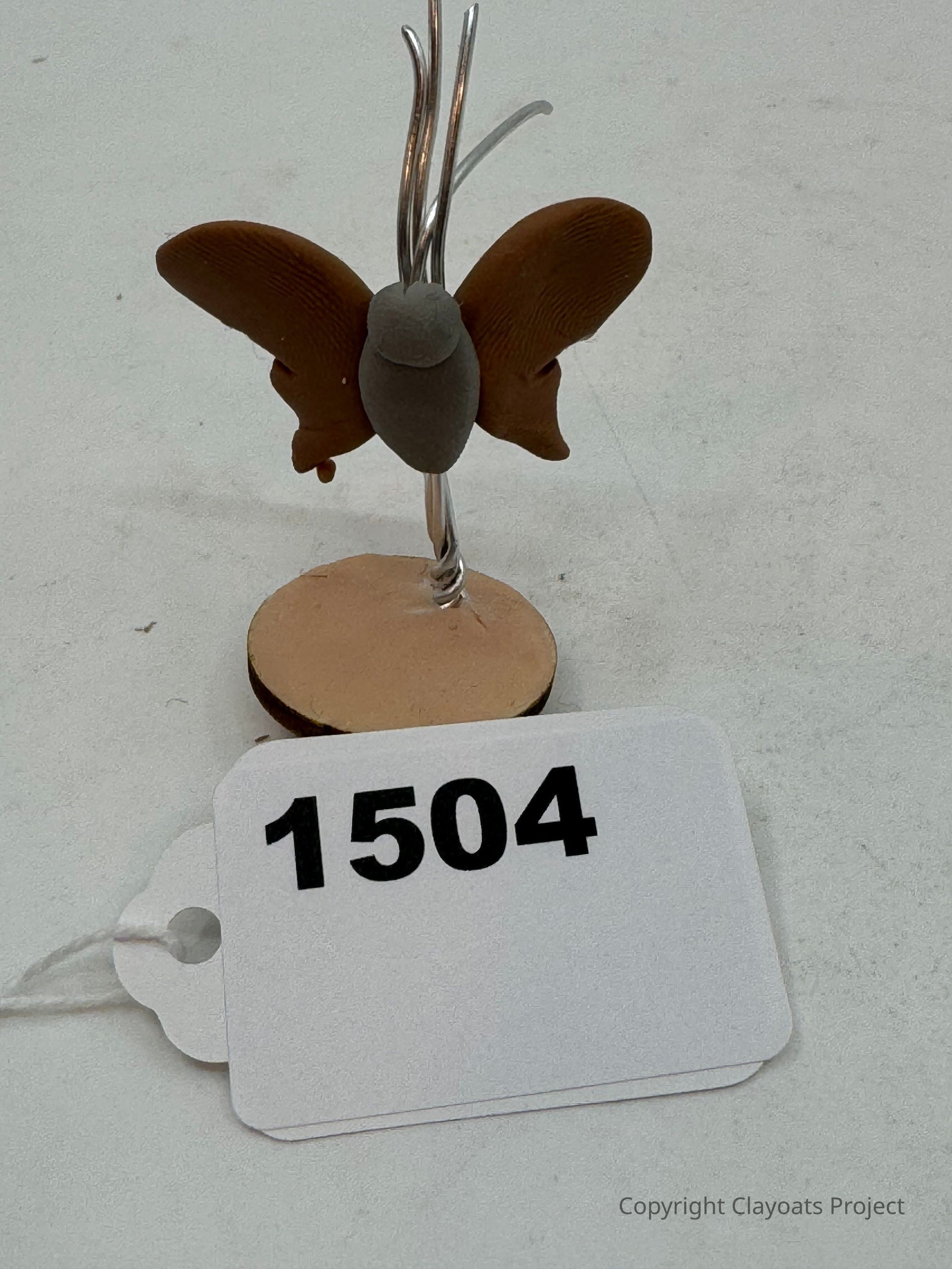 The image shows a small, handcrafted sculpture of a butterfly with brown wings and a gray body, all attached to a metal wire that extends upward from a circular beige base. This structure is displayed on a pale surface. The butterfly's wings have detailed texture, adding a lifelike appearance to the intricately crafted figure. The metal wire supports and elevates the butterfly above the base, providing an elegant contrast to