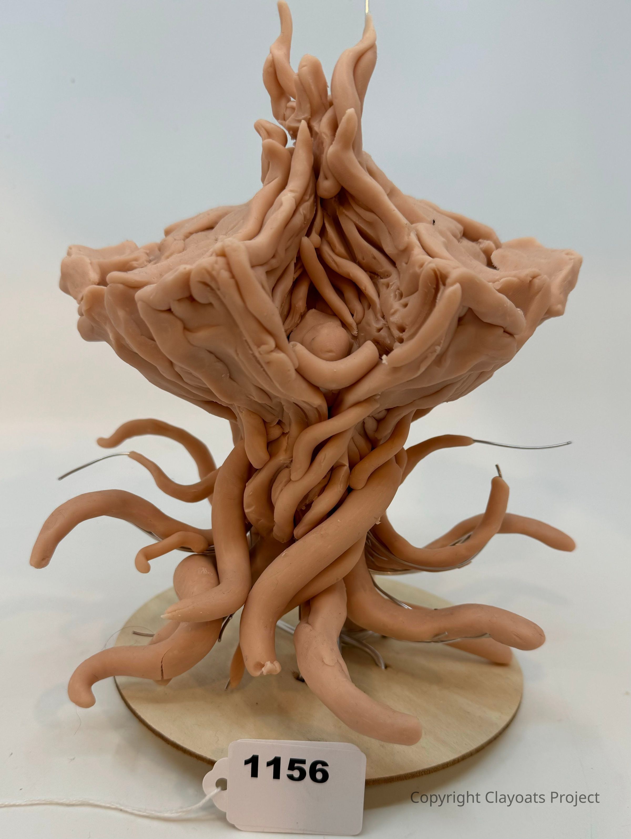 The image features a unique sculptural piece made of clay with intertwined, tentacle-like forms extending both upward and downward, resembling an abstract organic entity. It is set on a circular wooden base, lending stability to the dynamic, intricate design that suggests movement or growth. The clay is molded in swirling patterns, adding texture and visual interest, while the earthy tone of the clay contrasts subtly with the light-colored background.