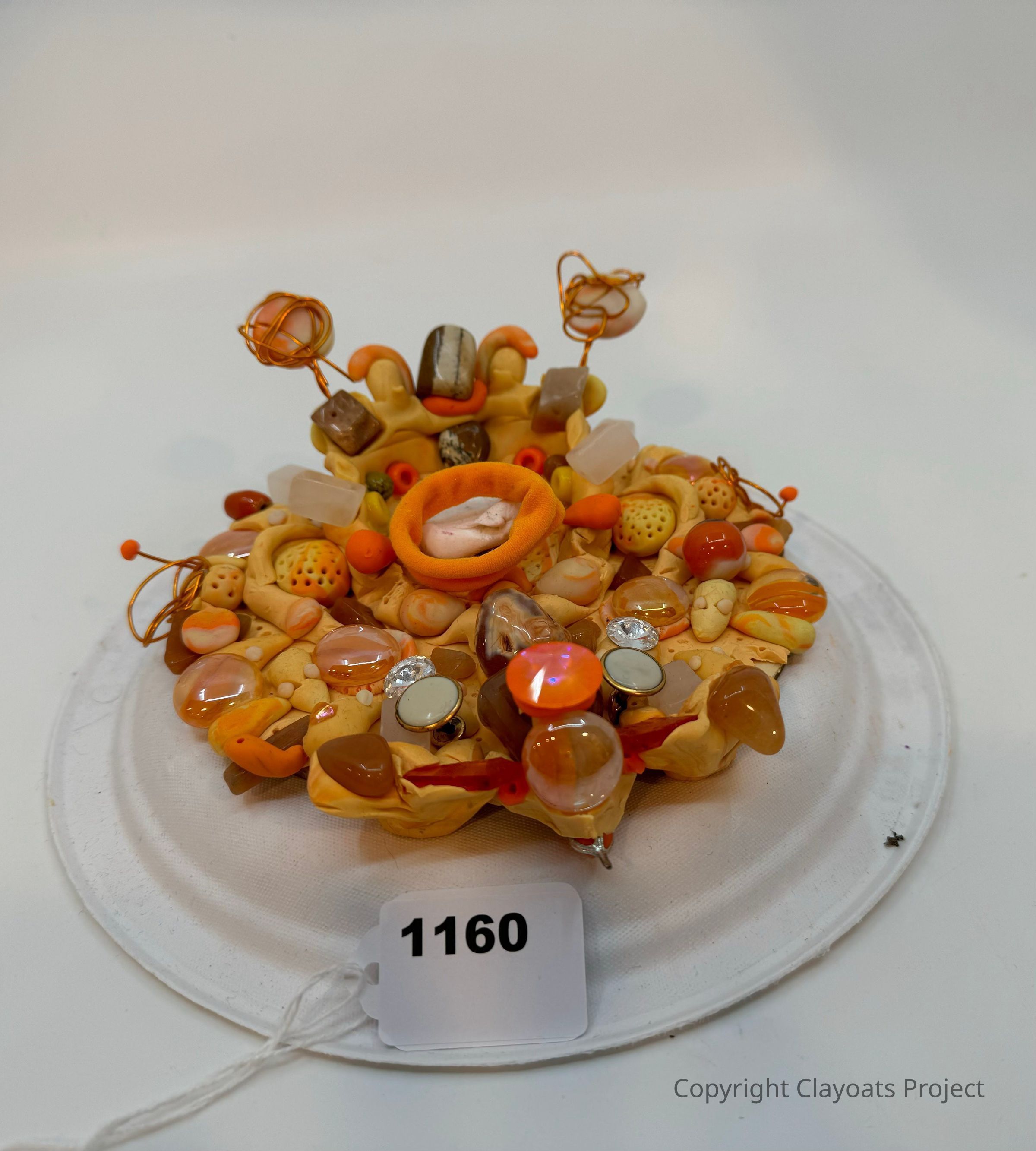The image displays a creative, multi-textured art piece composed of various small objects such as beads, gemstones, and wire ornaments, arranged on a circular white base l. The dominant colors are warm tones, including shades of orange, yellow, and brown, and there are reflective surfaces that suggest a whimsical, intricate design, potentially representative of abstract or imaginative artistic expression. The central feature resembles a soft, padded component, surrounded by an assortment of shapes and materials.