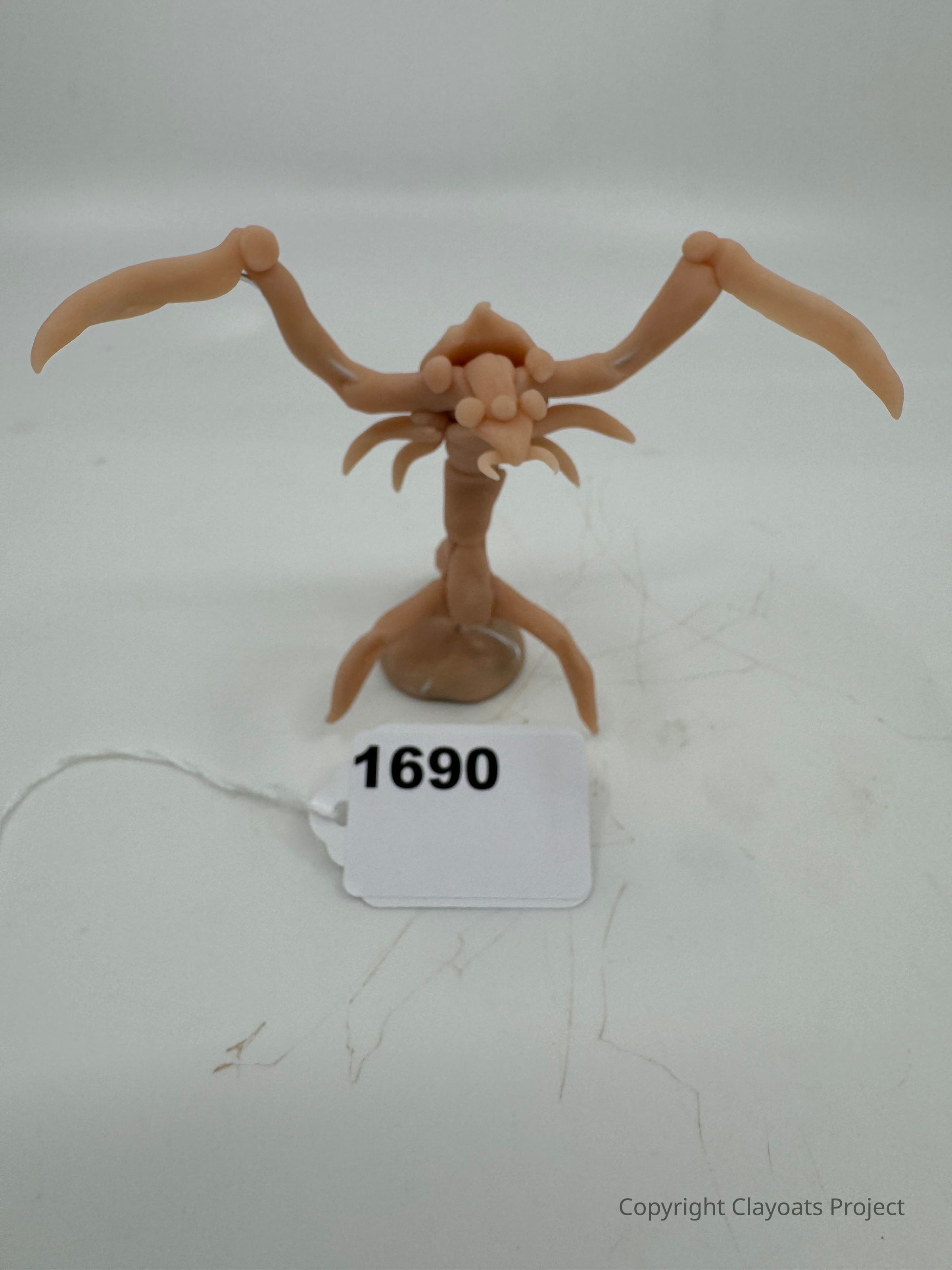The image shows a small, sculpted figure made of a light brown, clay-like material, with a unique and abstract form that resembles an alien or insect-like creature, featuring elongated limbs extended upward and outward, a body with multiple protrusions or appendages, and a circular base for support; below the figure.