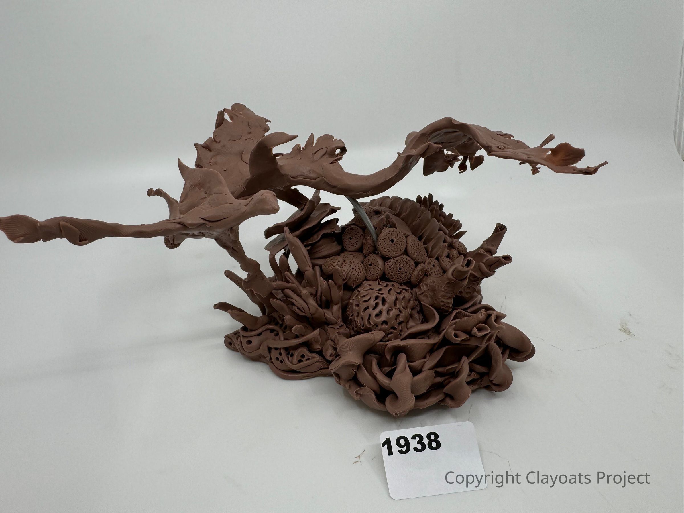 The image shows an intricate sculpture crafted from a brown material, likely clay, resembling abstract organic forms, such as leaves and coral-like structures, sprawling in various directions with a central cluster of round, textured shapes. The detailed and expressive design seems both chaotic and harmonious, representing a blend of natural and imaginative elements. The background is a simple, solid light color, which emphasizes the sculpture.