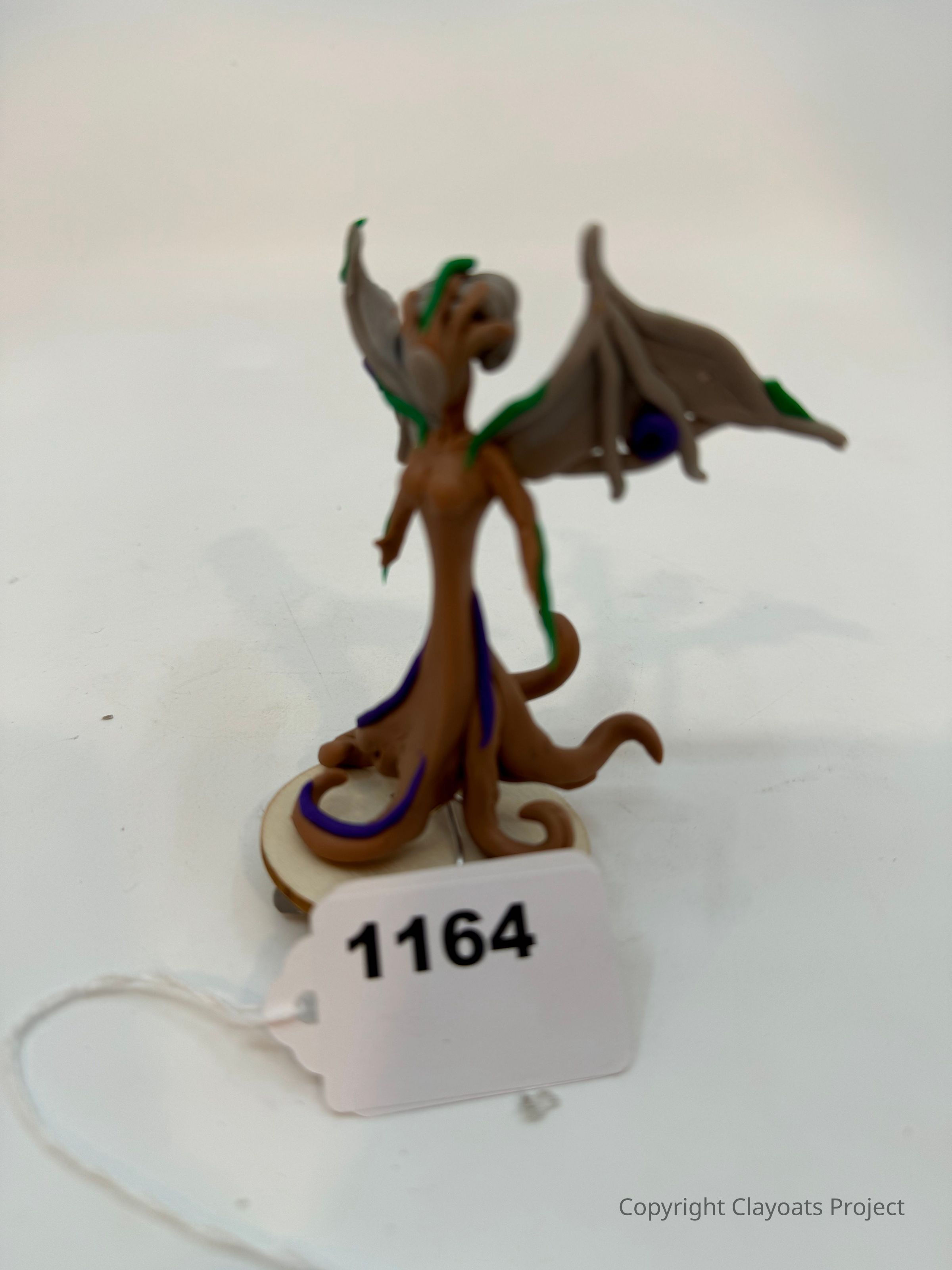 The image depicts a small, detailed figurine of a fantastical creature with elongated limbs and large, bat-like wings adorned with green accents and dark blue orbs, standing atop a circular base. The creature has a unique design, featuring a swirling brown body with hints of purple, creating a mystical and otherworldly appearance. Attached to the base.