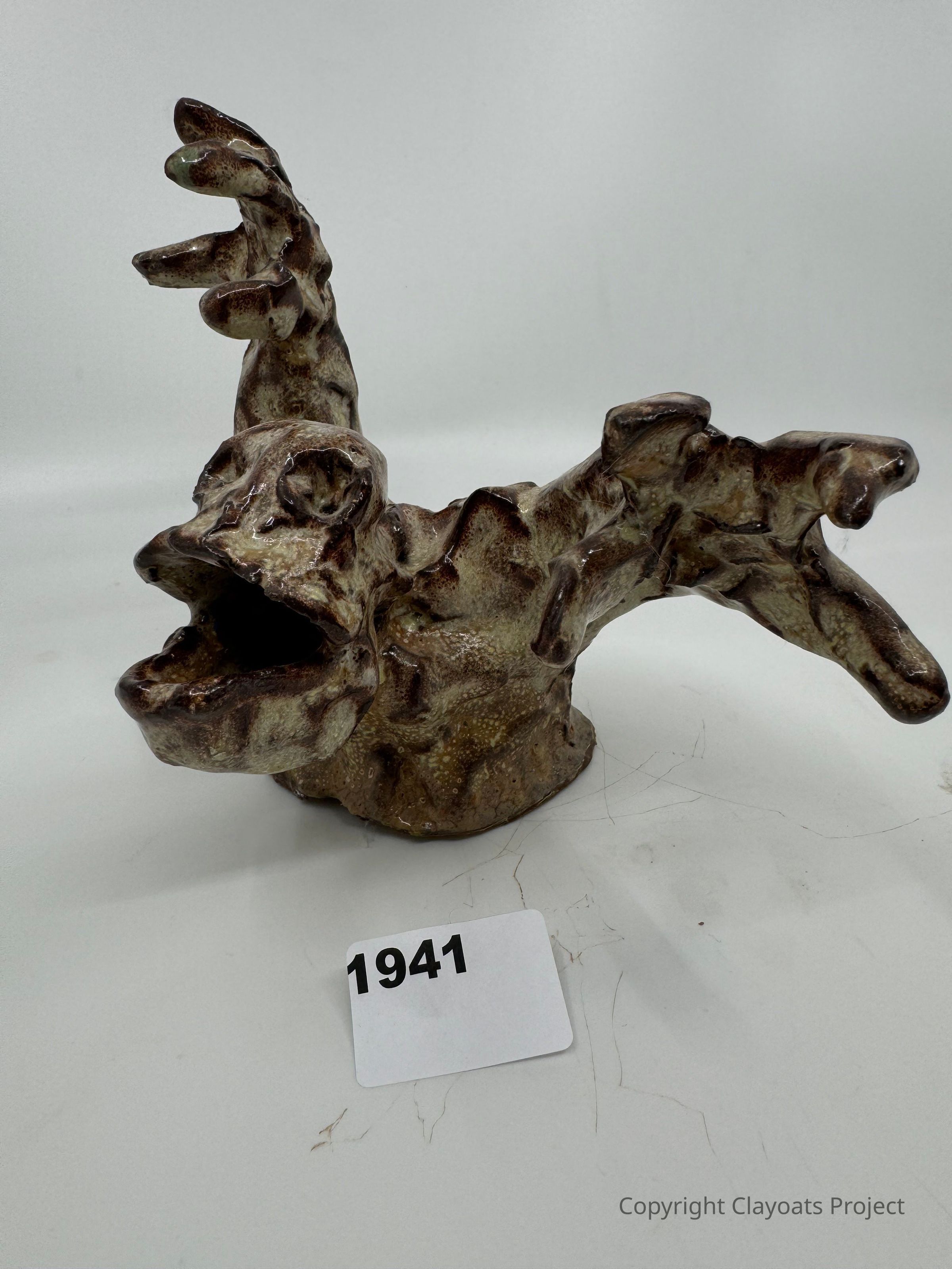 The image showcases a uniquely shaped, abstract ceramic sculpture featuring an expressive, organic form with a textured surface, displaying a rough, earthy finish with shades of brown and beige. The sculpture seems to depict a whimsical creature with an open, exaggerated mouth and protruding appendages, resembling limbs or antlers, curling and extending in dynamic, flowing motions.
