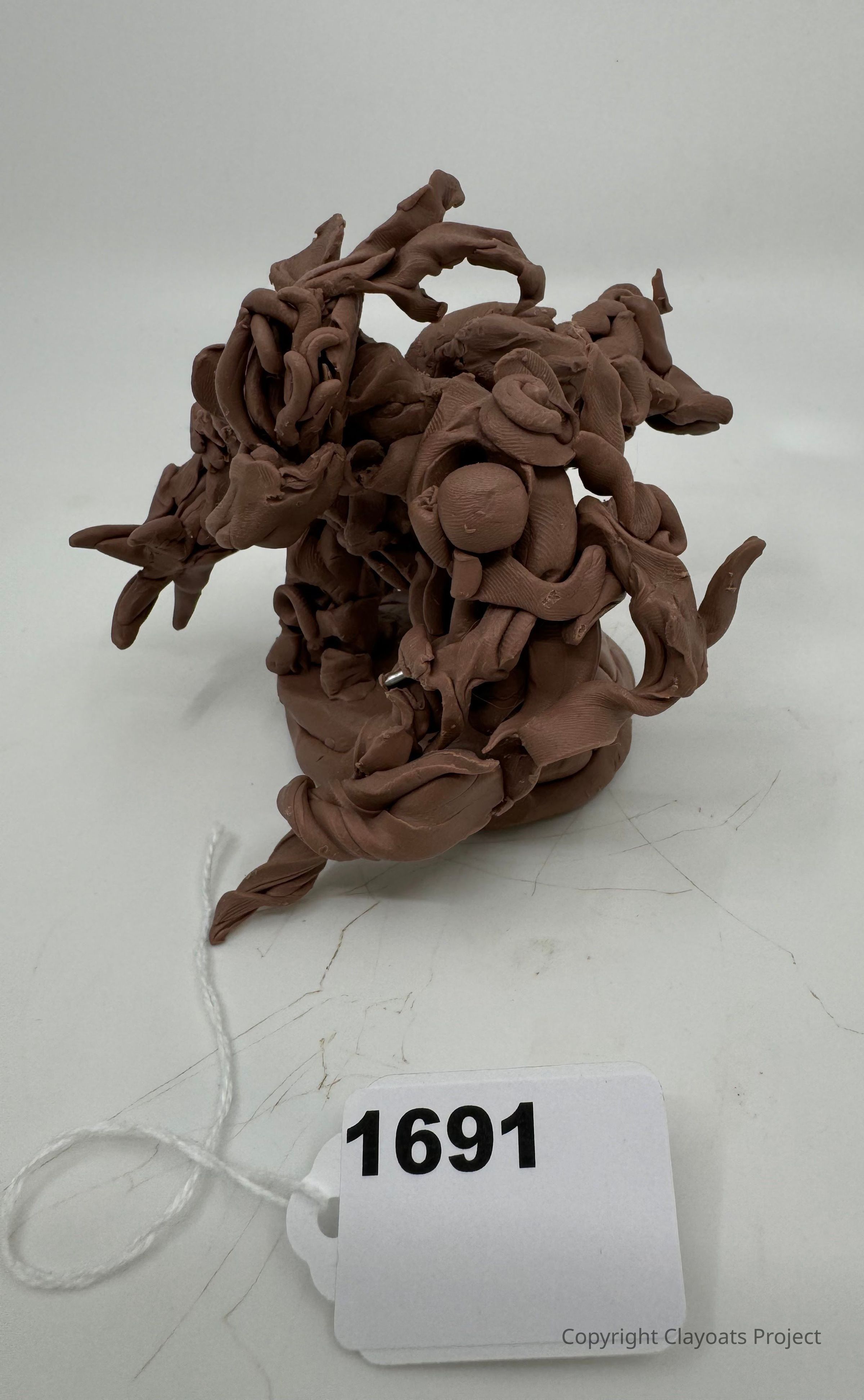 The image shows an intricate clay sculpture with swirling, organic forms, displaying a complex, abstract design characterized by sinuous lines and rounded shapes, all in a uniform earthy brown color. Its dynamic, flowing structure seems to twist and turn, suggesting movement and energy. The sculpture is placed on a plain, light surface.