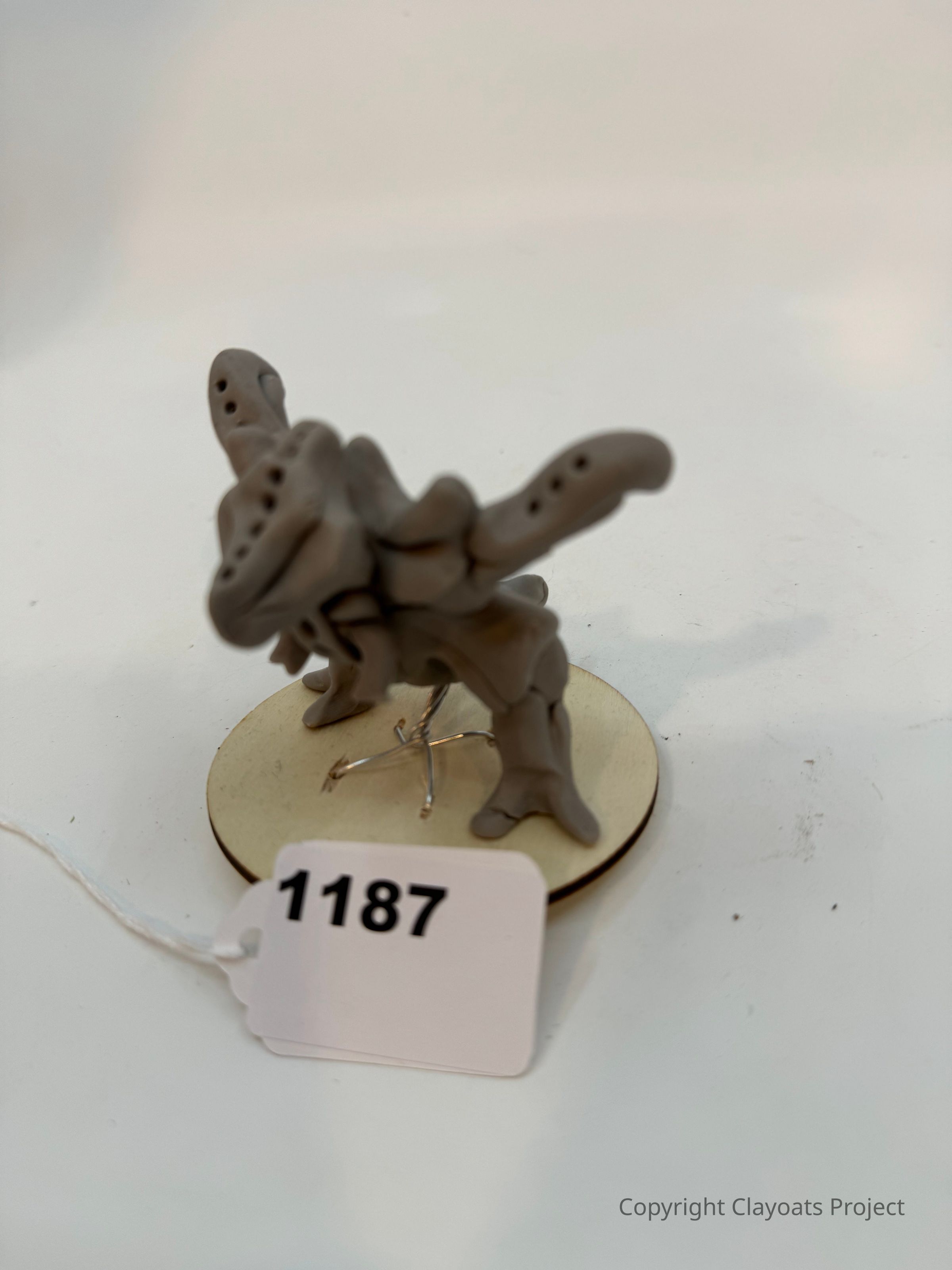 The image displays a small, abstract clay sculpture set upon a round wooden base, featuring various curving and protruding forms that resemble an imaginative creature or structure; the sculpture is held up by a thin wire positioned against a neutral white background that emphasizes the details and shadows of the sculpture, highlighting its unique and creative design elements.