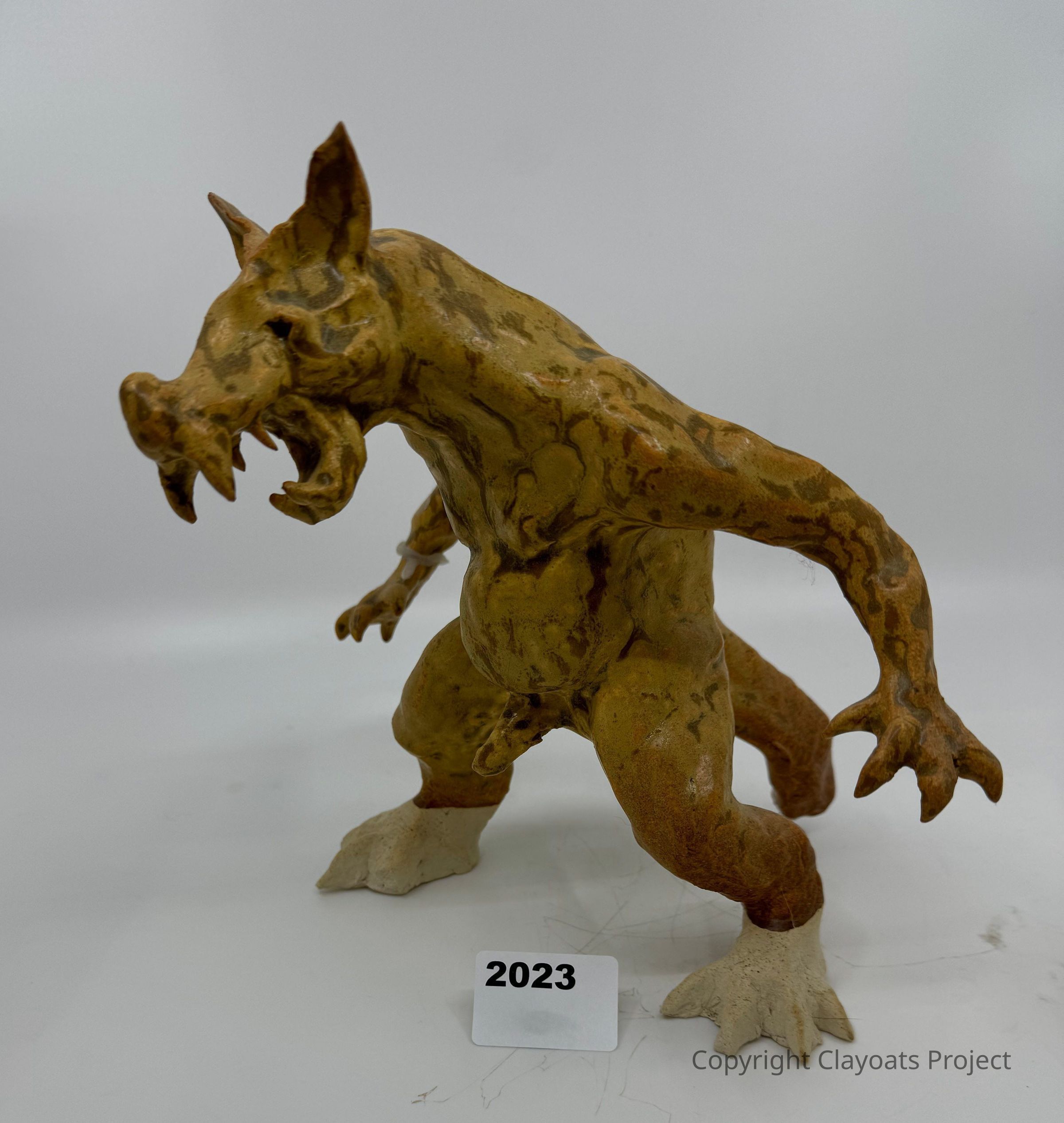 The image shows a detailed sculpture of a mythical creature resembling a werewolf, standing on a white surface with an aggressive posture, its mouth open and sharp teeth visible, with pointed ears and textured fur that appears carved or molded; the creature has muscular limbs ending in clawed hands and feet, with the entire figure painted in earthy tones that enhance its fierce and intimidating presence, and a small rectangular card with the number "2023" placed in front of its feet on the surface, possibly indicating the year