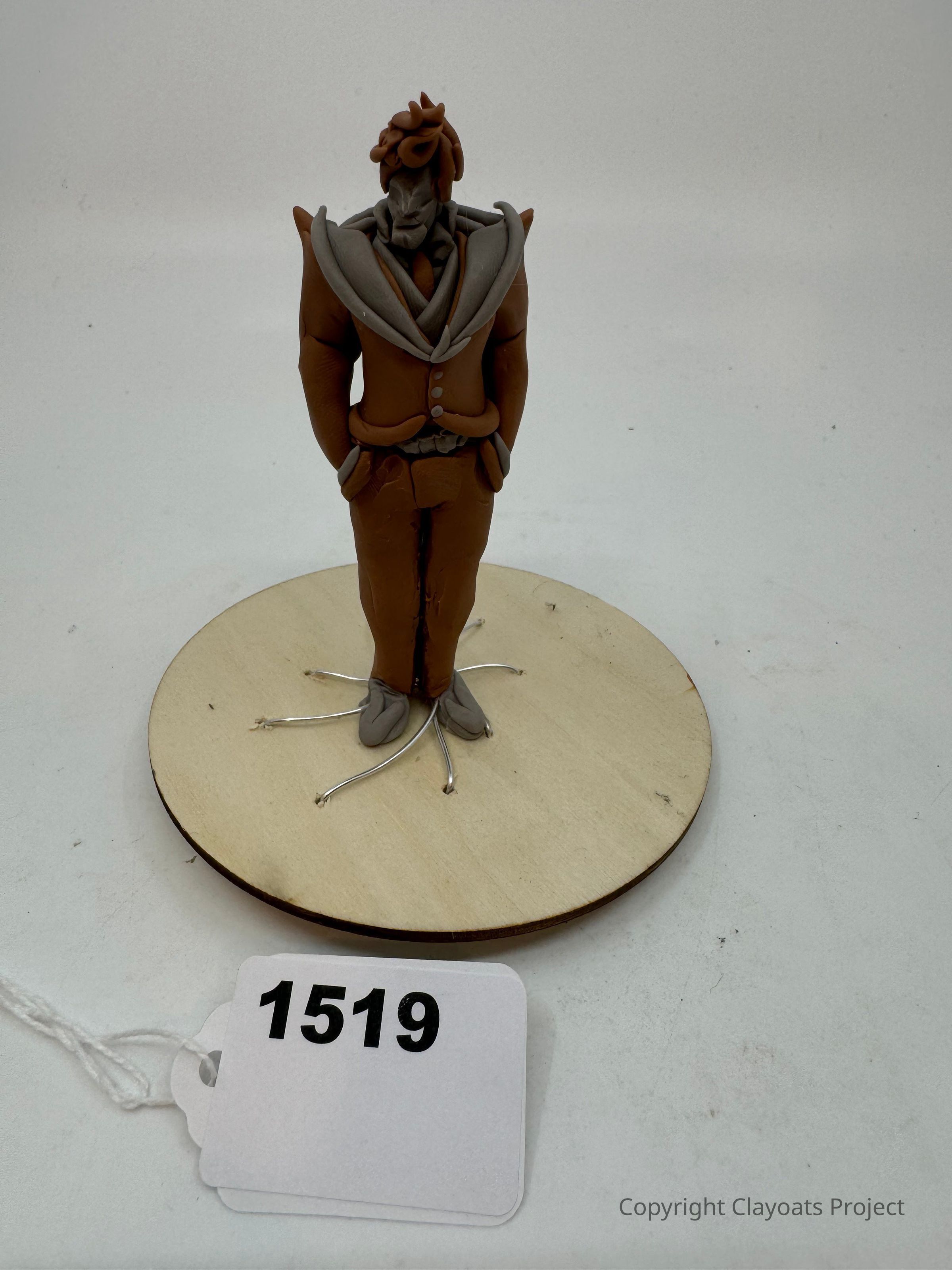 The image showcases a clay figure of a man in a brown suit with a grey-collared jacket and matching grey shoes, standing on a round wooden base. The figure has distinctive, stylized hair and a serious expression. Metal wires are visible extending from the feet, giving an impression of roots or supports. In front of the base.