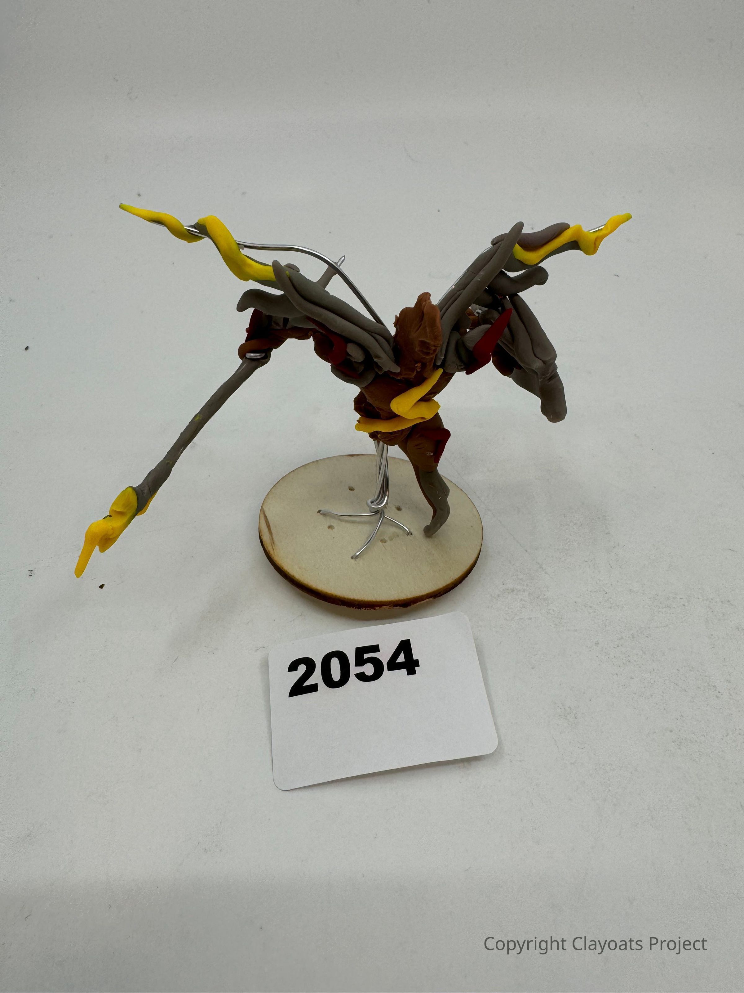 The image shows a creatively designed figurine with multiple arms that appear to be made from gray and yellow clay, featuring intricate details, including elongated appendages extending outward. The background is a plain white surface, which helps emphasize the unique details and colors of the figurine, showcasing its dynamic and artistic composition, likely indicative of a fictional or fantasy character.