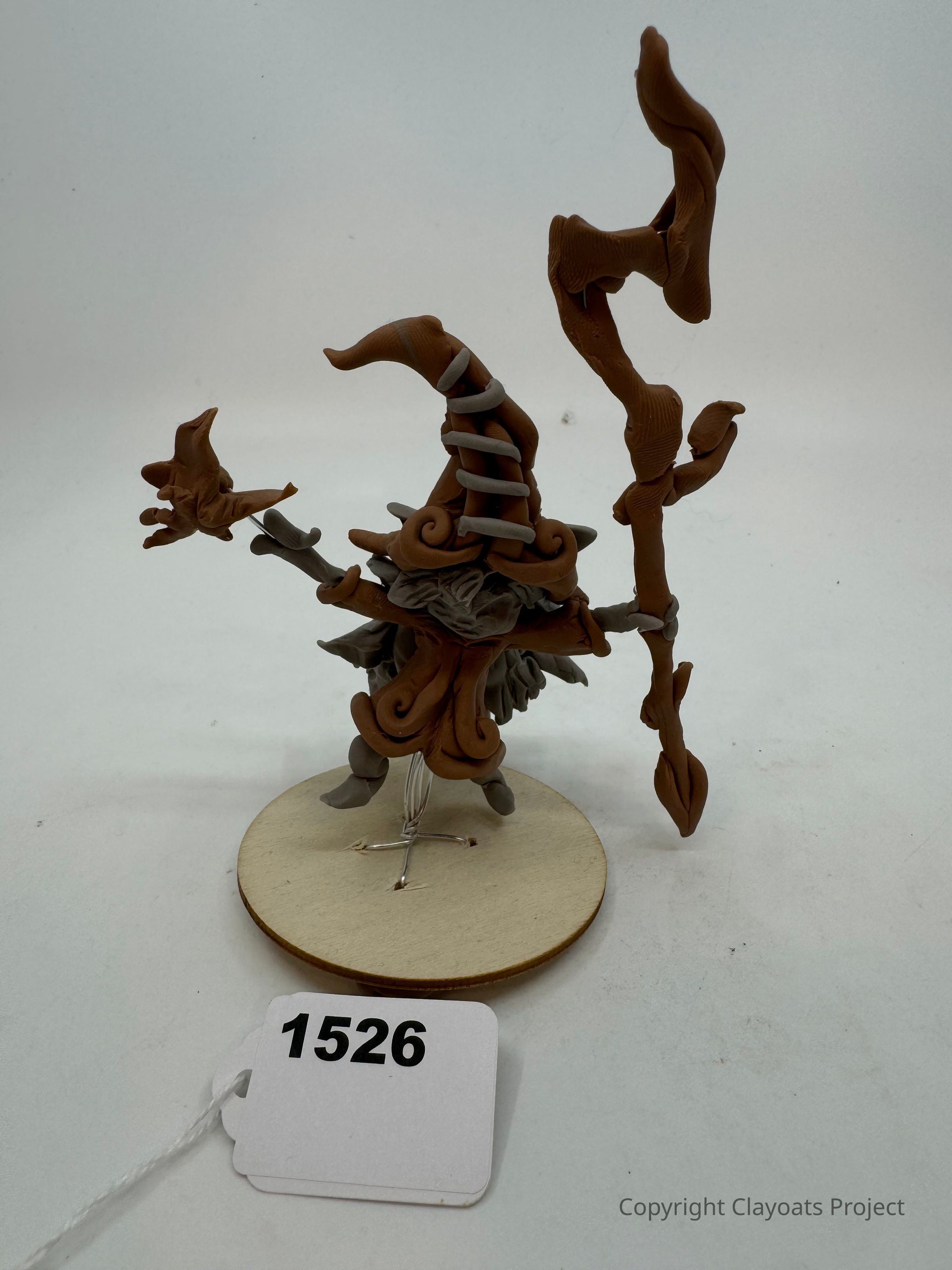 The image shows a whimsical figurine crafted with earthy-toned clay, featuring a wizard-like character with a tall, spiral hat and intricate details, such as a curled beard and layered robes, standing on a round wooden base while holding a gnarled staff in one hand and a star-tipped wand in the other, exuding a sense of fantasy and creativity.