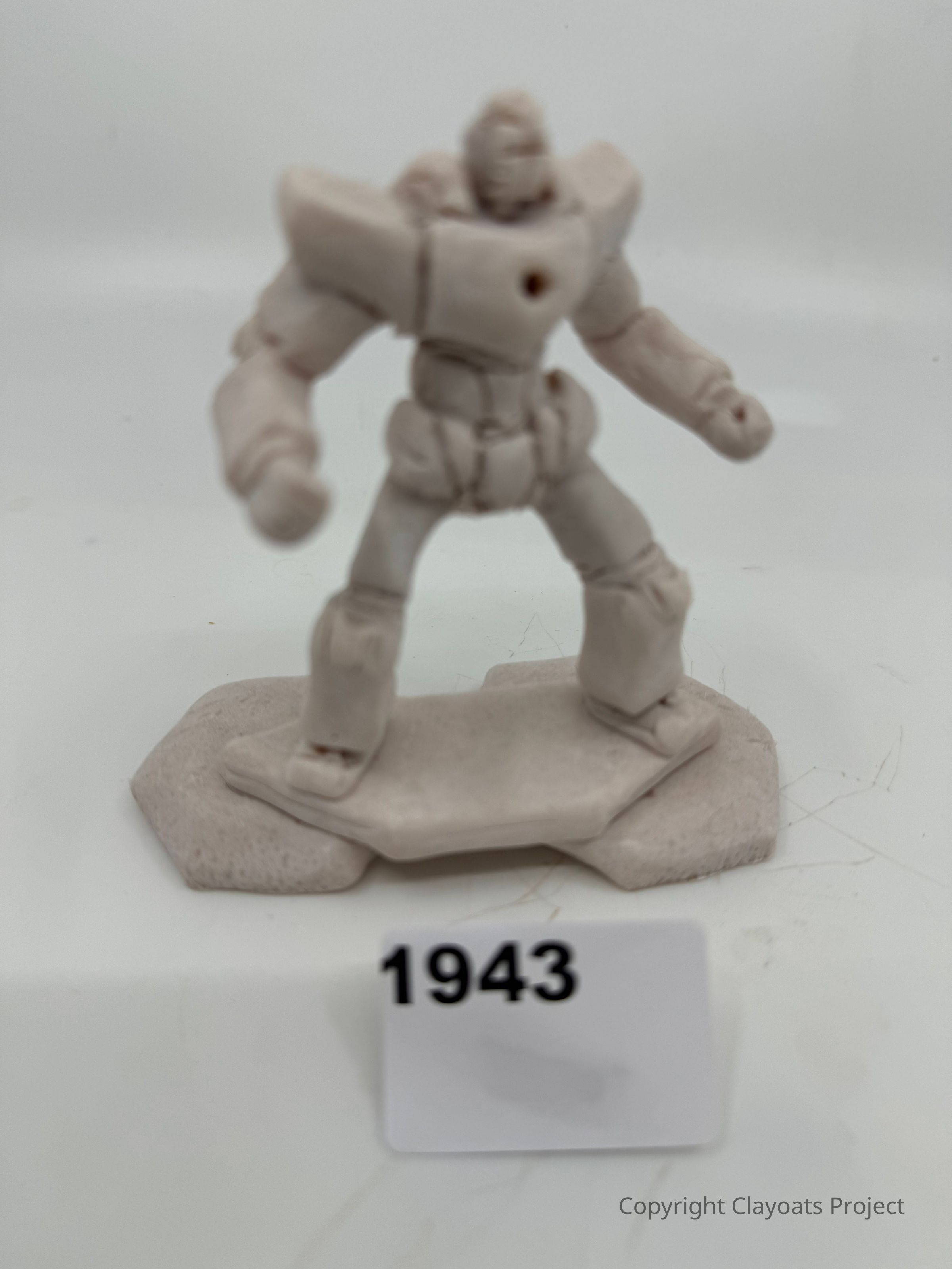The image depicts a small, pale, clay-like figurine of a robot standing on a simple base. The robot is designed with broad shoulders and articulated limbs, suggesting a humanoid form. The figurine’s texture appears rough, indicating an unfinished or handcrafted quality. Its stance is dynamic, with slightly bent knees and clenched fists, giving it an energetic appearance. The background is plain and white, focusing attention solely on the figurine, which