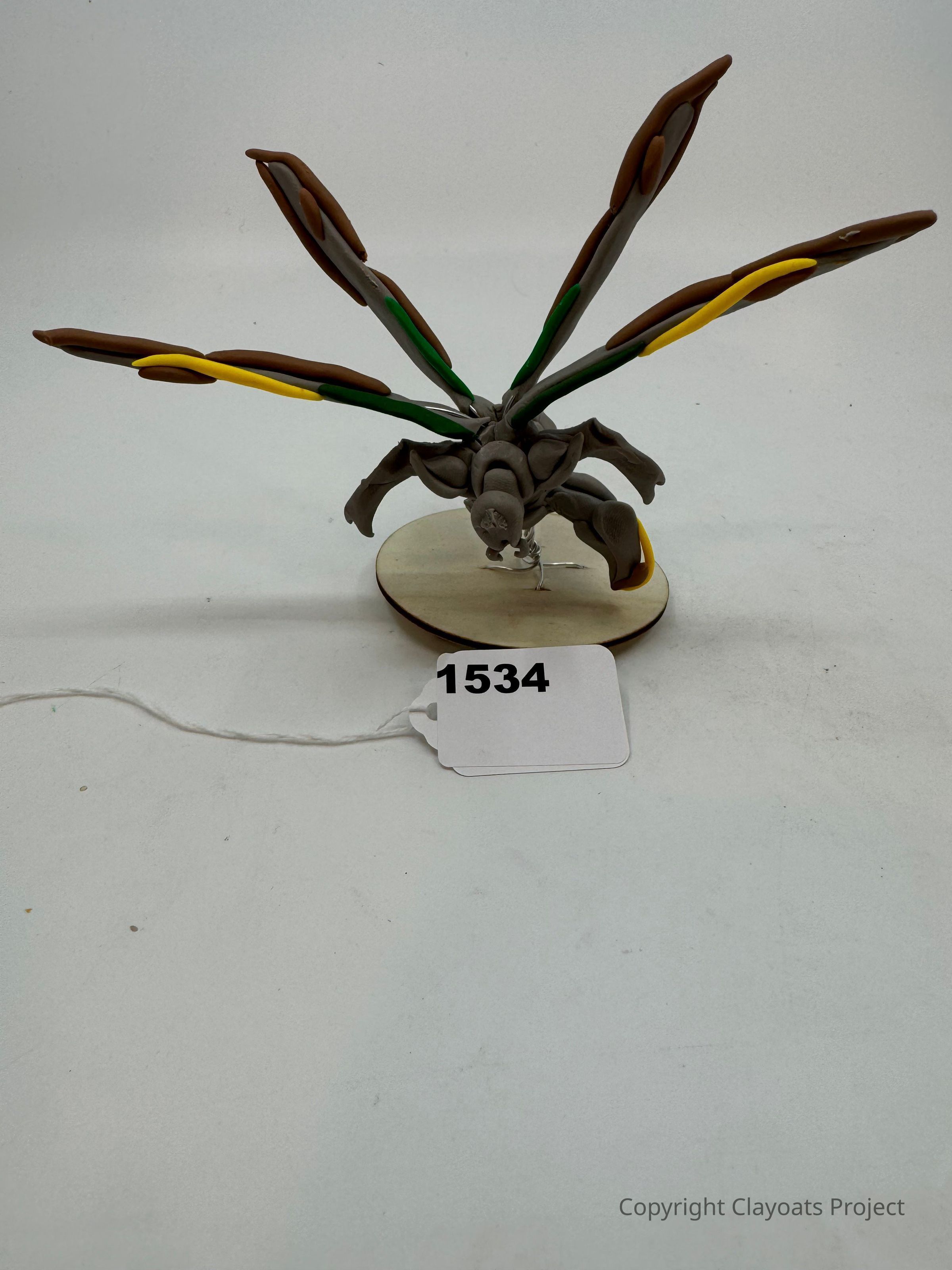 The image depicts a sculpture or model situated on a circular base, featuring an intricate design with six elongated, wing-like protrusions radiating outward in various directions, including colors like brown, yellow, and green. The central body of the structure resembles a creature with complex, abstract anatomical features, emphasizing an imaginative, possibly extraterrestrial or insect-like form. The piece is carefully balanced on a flat surface.