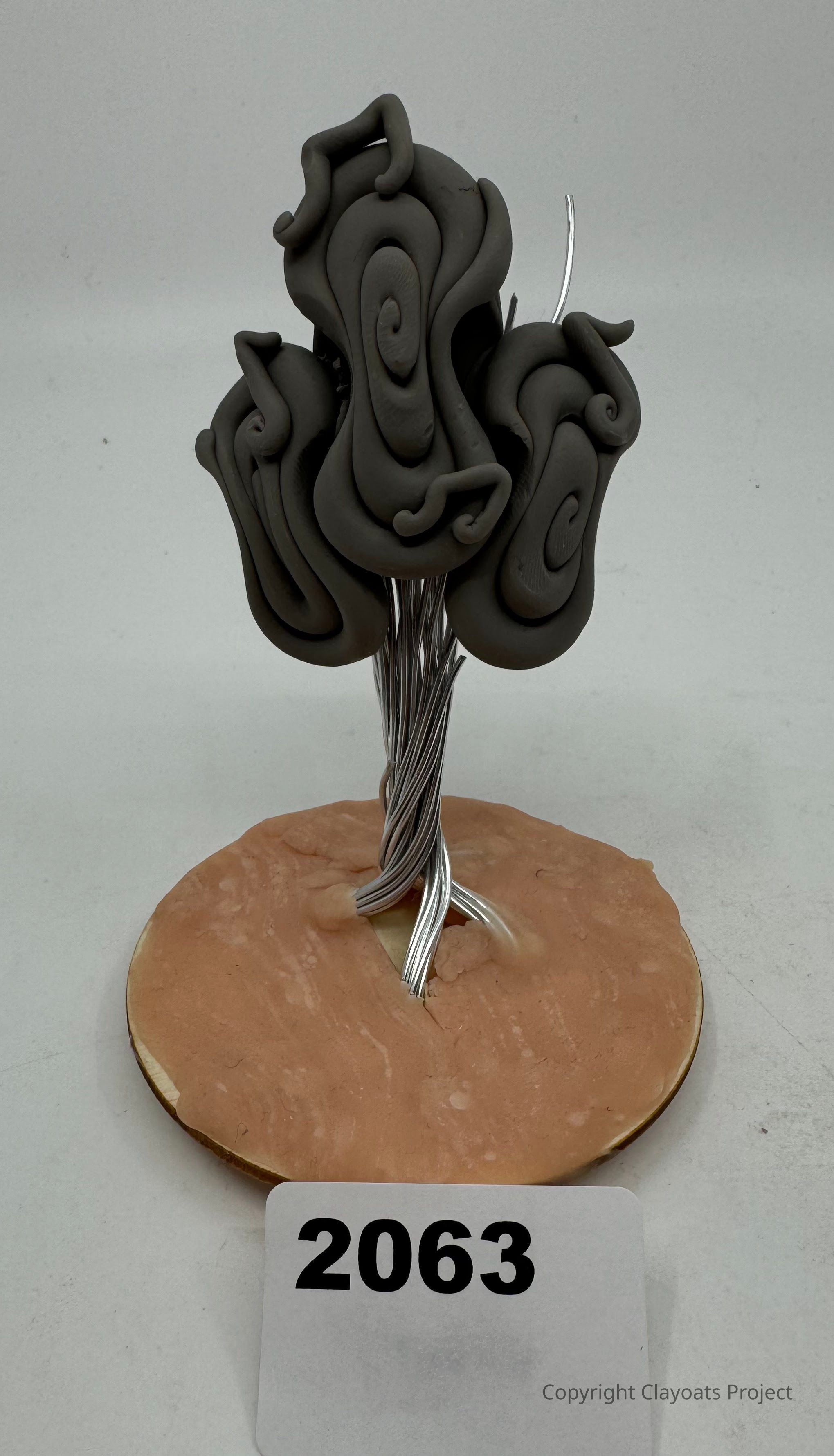 The image shows an artistic sculpture resembling a tree with twisted, swirling dark branches made of a smooth material, like clay or resin, standing on a circular, flat base that appears to be a light brown color. The tree’s trunk is fashioned from silver wires, intricately intertwined to create a supportive structure that ascends into the abstract branches above. The whole piece is placed against a plain background.