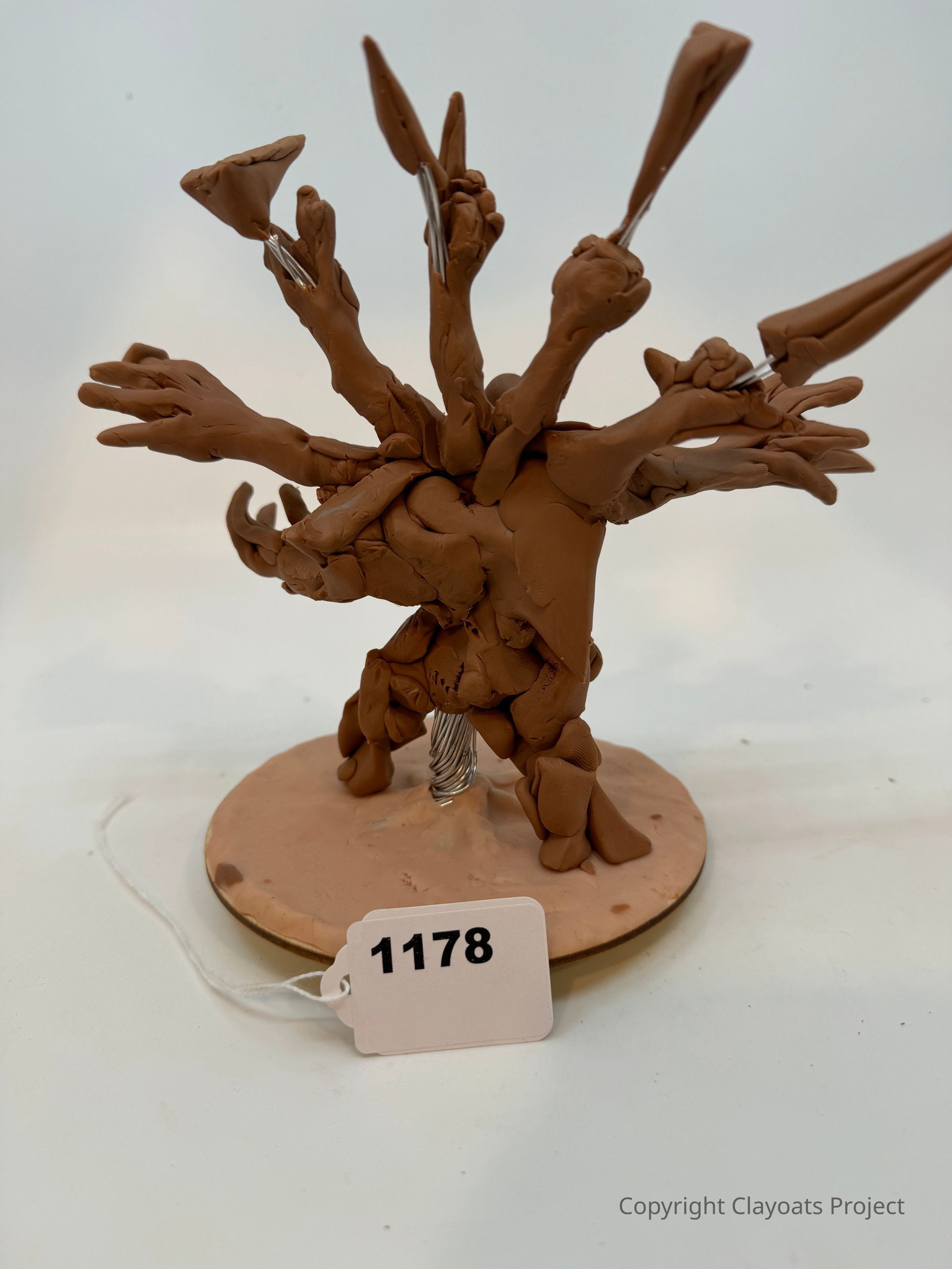 The image depicts a sculptural creation made from brown clay, featuring a central form with multiple elongated appendages, resembling a fantasy creature; it is mounted on a circular base that has a wire support visible underneath, , resting on a smooth, light-colored surface, showcasing the intricate and imaginative craftsmanship involved in its construction, possibly intended for artistic or educational display.