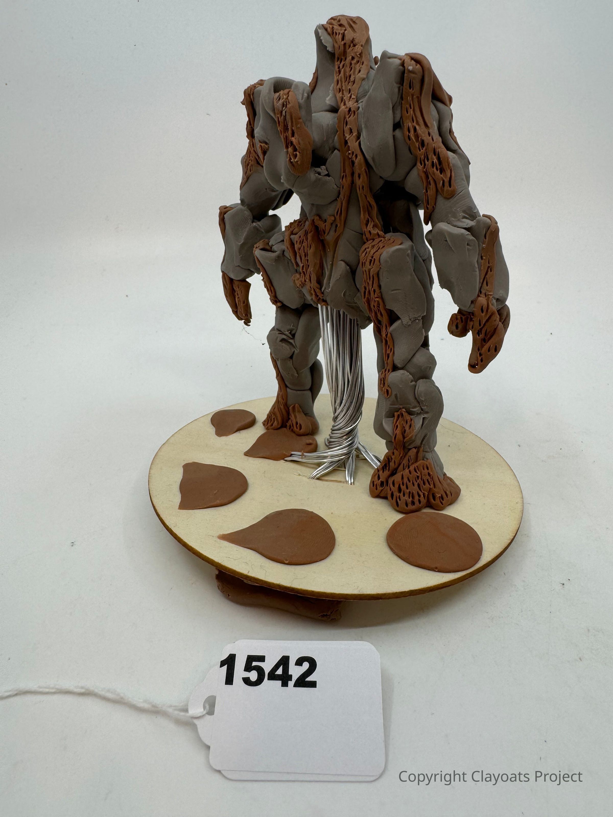The image shows a sculpture of a humanoid figure made from gray and brown clay, mounted on a circular base with clay leaves and wire details. The figure appears abstract with a fragmented surface texture resembling rocks or bark. The background is plain white, enhancing focus on the artistic creation. The piece stands tall with bulky limbs, giving an impression of strength and stability, while the base provides an earthy contrast.
