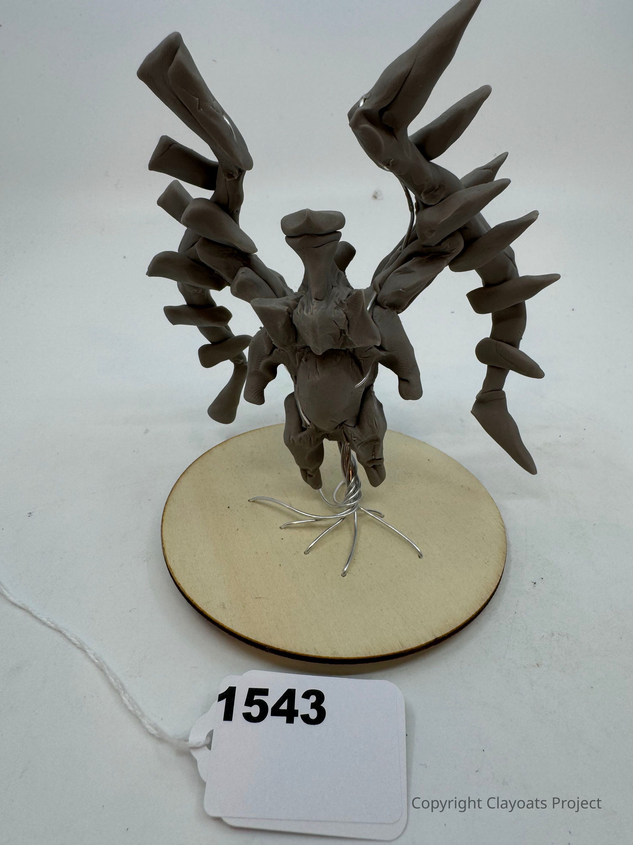 The image showcases a unique sculpture crafted from dark gray clay, depicting a fantastical creature with multiple angular wings and a humanoid form. It stands upright on a circular wooden base, supported by a wire structure that extends outwards resembling legs or roots. . The background is a simple, neutral off-white color, emphasizing the central focus on