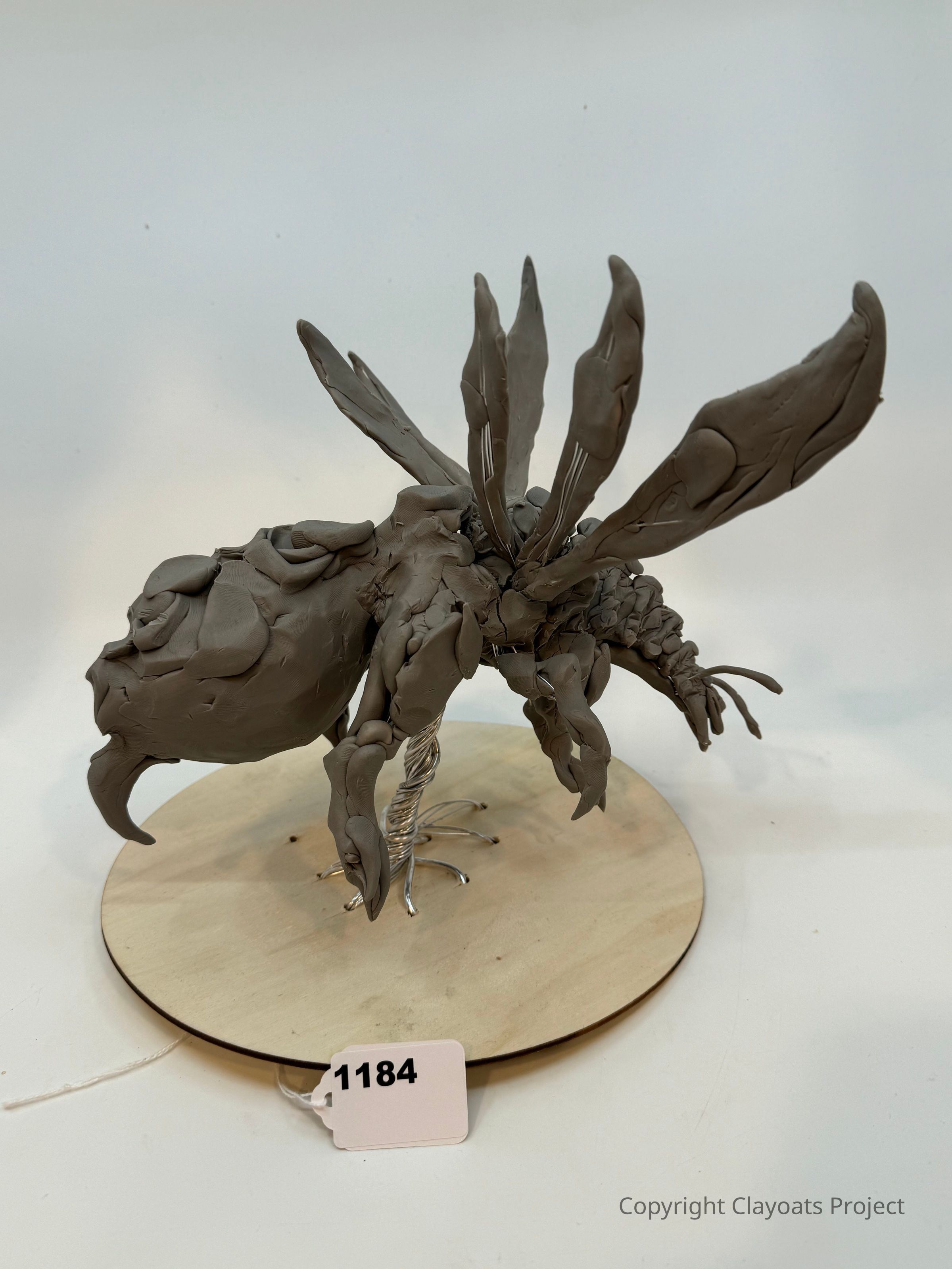 The image features a clay sculpture of a fantastical creature, resembling an insect with large, elaborate wings and a sturdy body, positioned on a round wooden base. Its limbs are intricately modeled, showcasing detailed, curved forms, while the wings stand prominently overhead. The entire structure is supported by a coiled, silver metallic wire underneath, which seems to be both the foundation and part of its artistic composition.