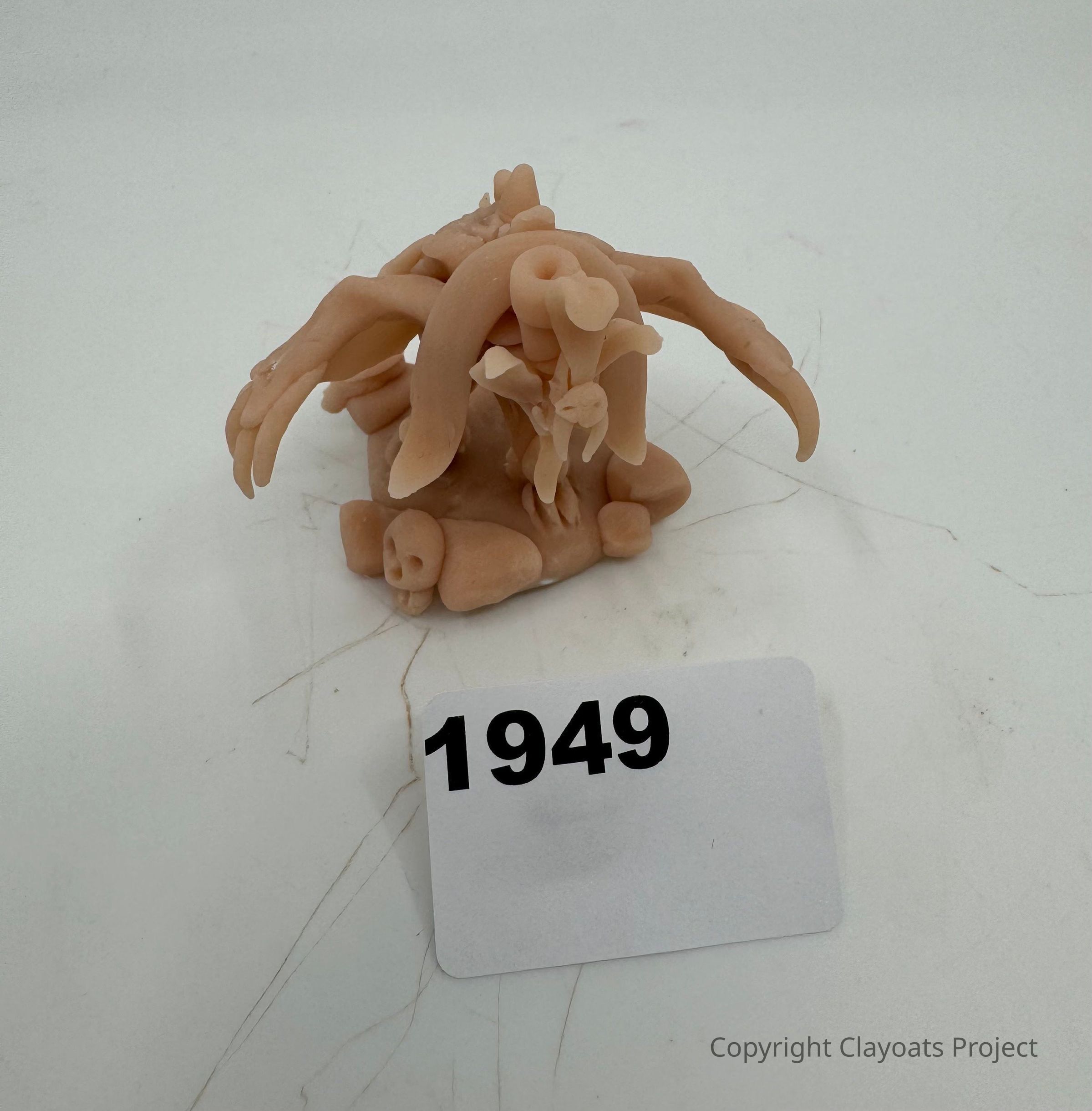 The image shows a small, intricate sculpture made from a flesh-colored material resembling plastic or wax, featuring twisted and abstract shapes that could be interpreted as organic or skeletal forms, like arms or tentacles, with a prominent circular hollow near the top. The background is blank and uncluttered, emphasizing the bizarre and complex structure at the center
