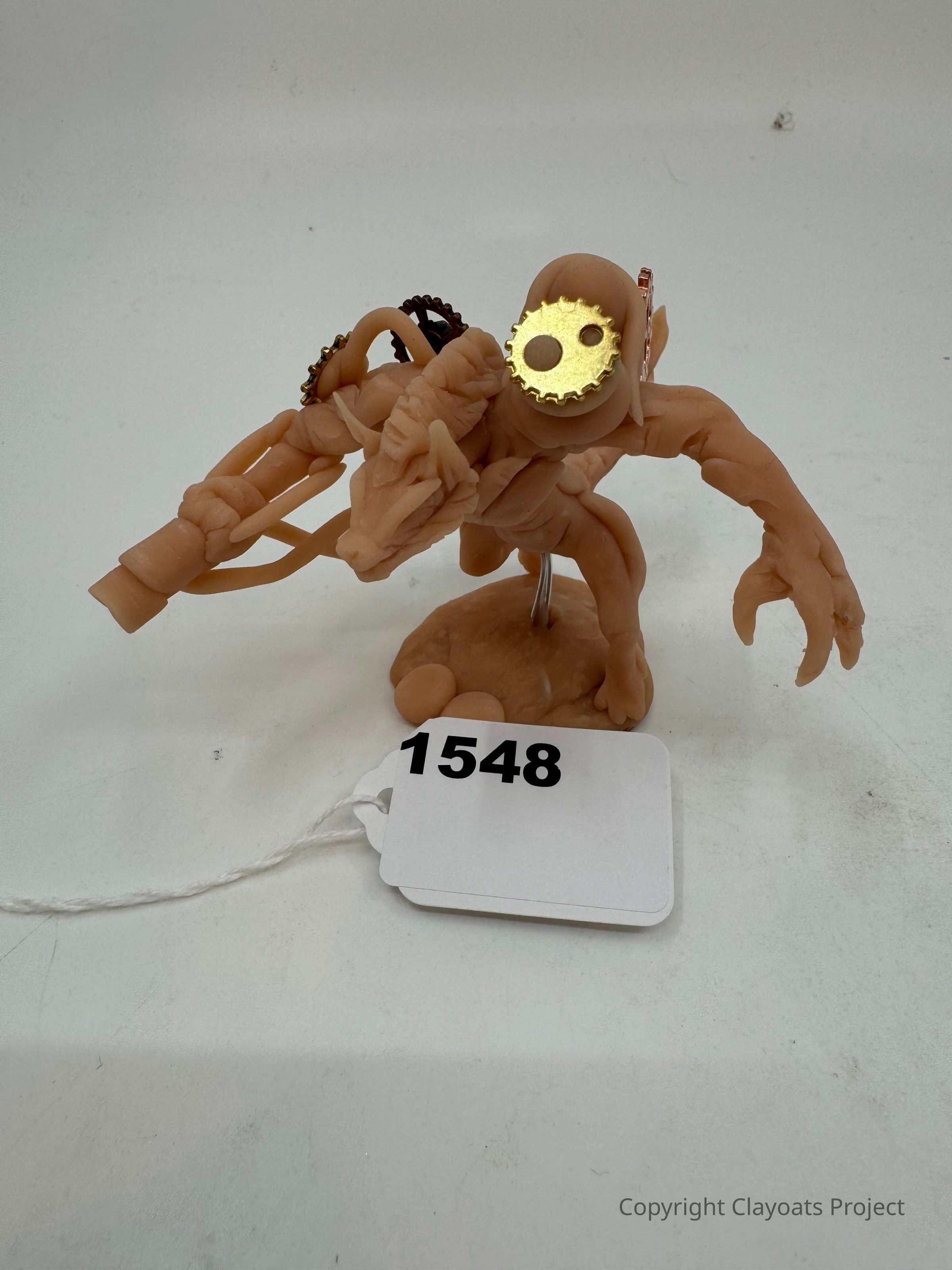 The image shows a small, detailed sculpture of a creature with elongated arms and a mechanical appearance, featuring a gold-colored gear incorporated into its design. It is mounted on a base, The sculpture's intricate textures and shapes suggest a blend of organic and mechanical elements, possibly representing a fantastical or science fiction-inspired character. The entire piece is positioned against a plain, light background, highlighting its distinct features and craftsmanship.