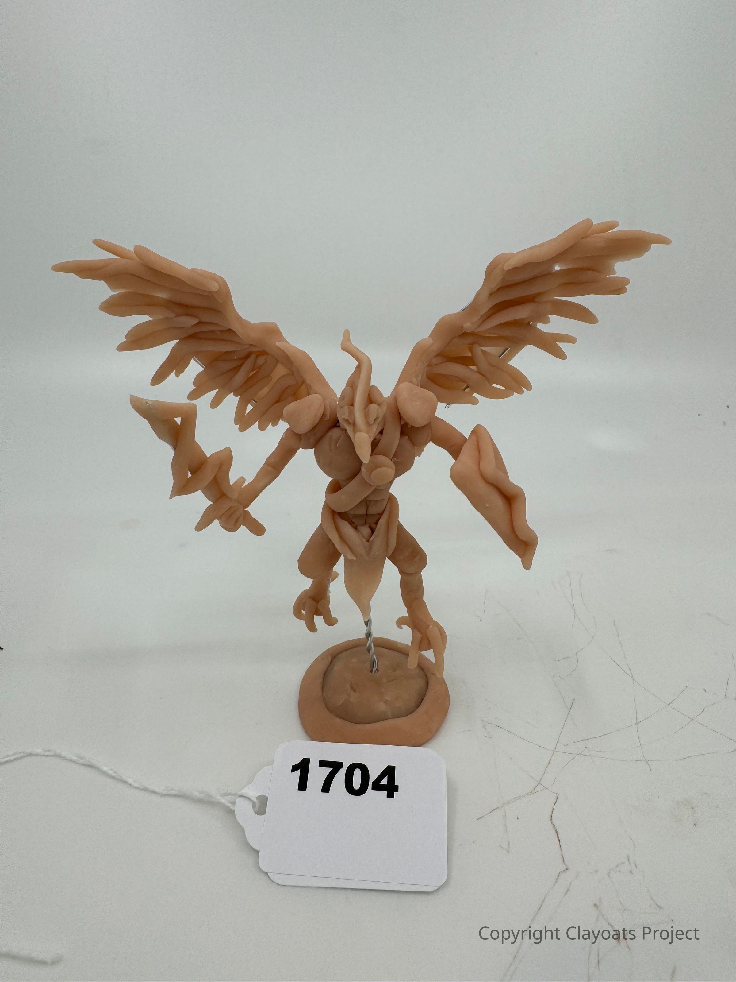 The image shows a unique clay sculpture of a creature with large, intricate wings, elongated limbs, and a distinctive body shape, standing on a small base, with a label numbered 1704 placed in front. The sculpture appears artistically crafted, with detailed attention to the wings and the overall posture, suggesting creativity and imagination. The pale, uniform color of the clay contrasts against the plain background, highlighting the figure's form and features, while a string is attached to the label, indicating it might be