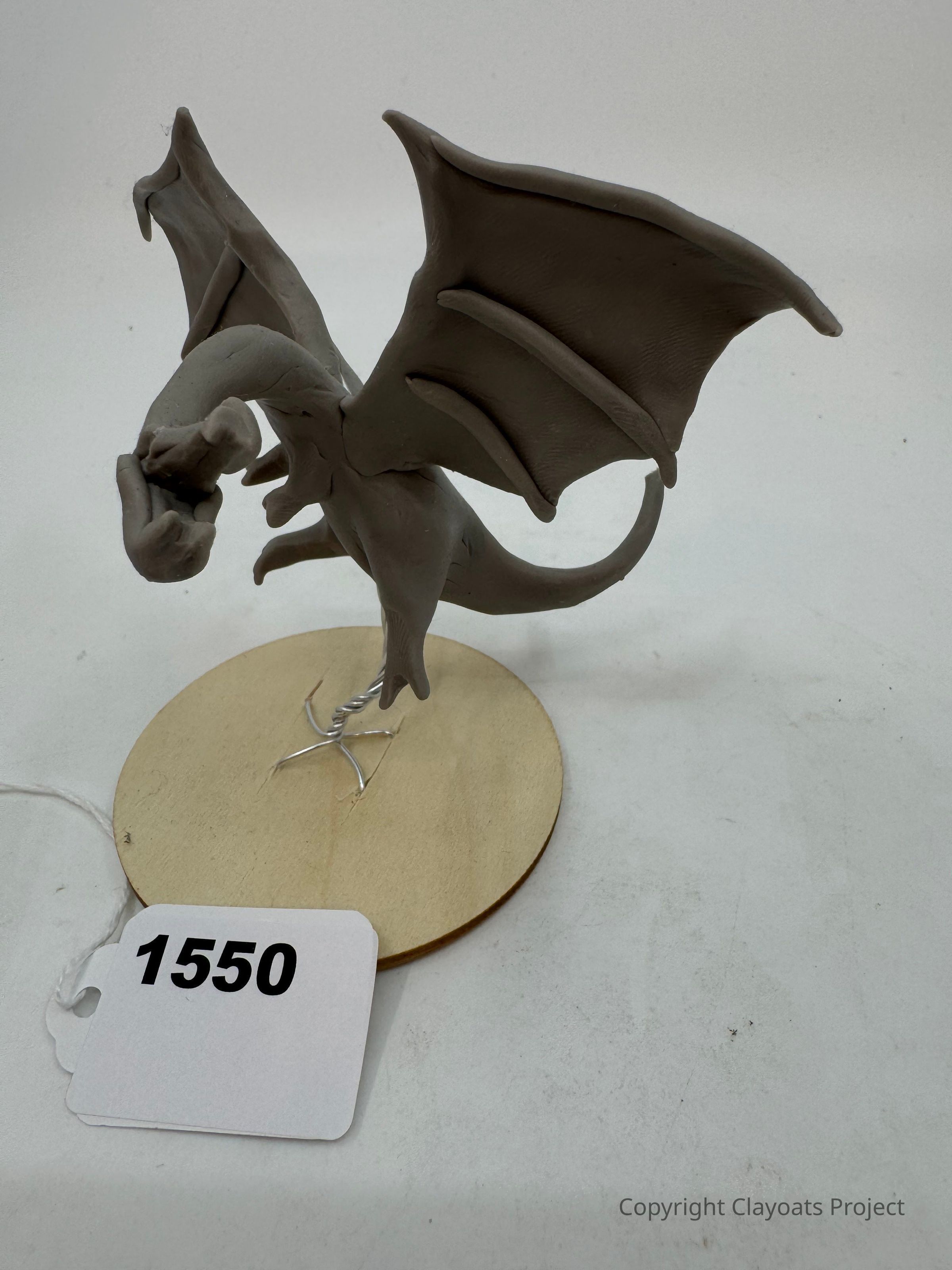The image showcases a detailed gray clay sculpture of a dragon with outstretched wings, perched on a wooden base with visible wire supports; the dragon, appearing dynamic and lifelike, holds a small object in its mouth The sculpture's intricate features, including its wings and body posture, convey a sense of realism and artistry, capturing the mythical essence of the dragon.