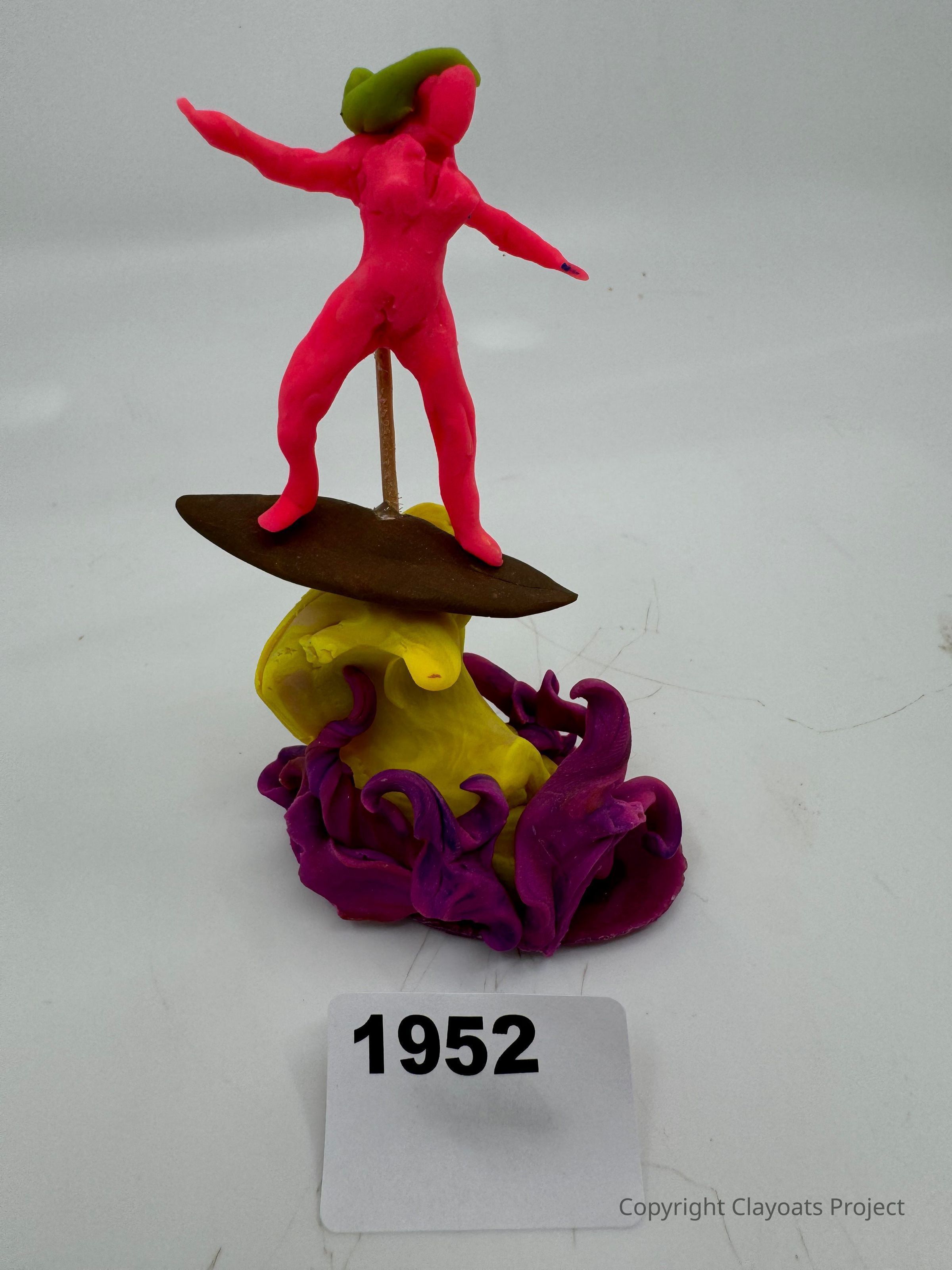 The image showcases a colorful clay sculpture featuring a pink human figure with a green hair piece balancing on a surfboard. The surfboard is mounted on a swirling yellow and purple wave-like base, creating a dynamic and vibrant appearance. t. The combination of bright colors and the surfing theme gives the piece a playful and creative vibe, highlighting the artistry involved in its intricate design and construction