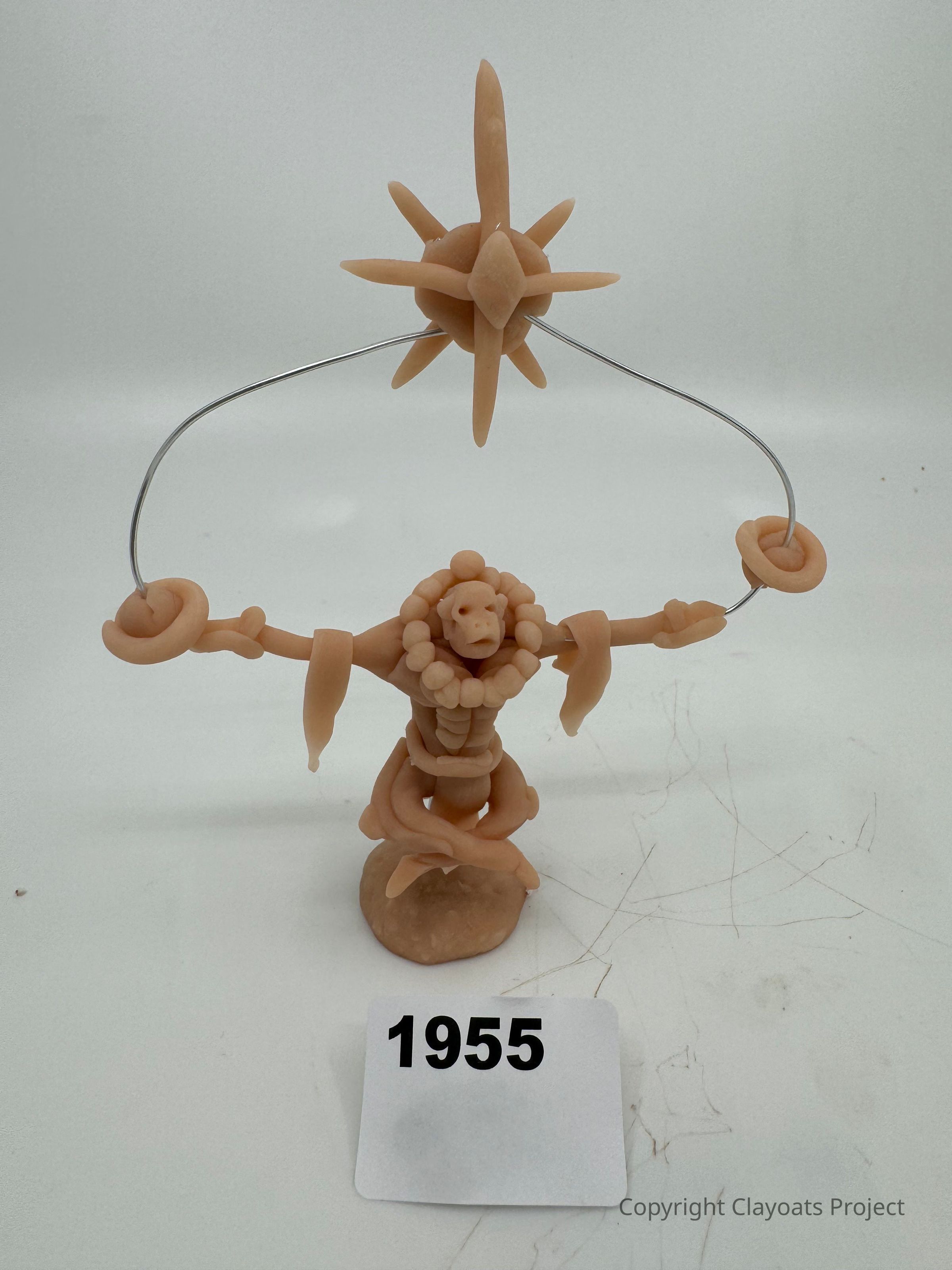 The image depicts a clay sculpture featuring a humanoid figure with raised arms connected to a wire, with a star-like object perched on the wire above. The figure has detailed features, including coiled and rounded shapes that form its limbs and body. At the base, the sculpture stands on a rounded platform, while its upper part is adorned with circular shapes. this