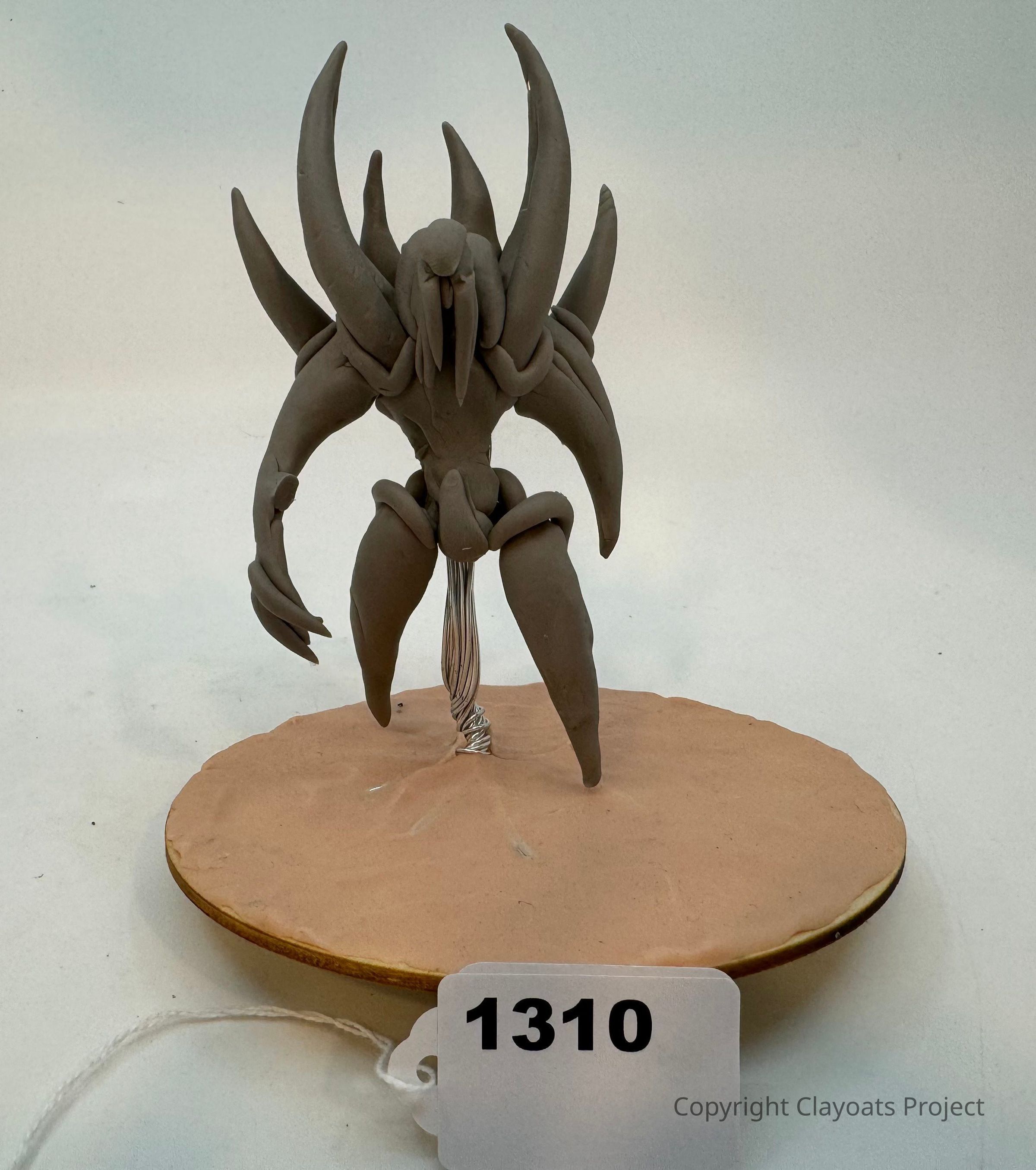 The image depicts a clay sculpture of a creature with multiple curved, elongated spikes protruding from its back and a humanoid form, standing on a circular, sandy base supported by a wire structure beneath it; the creature's imposing form, with claw-like hands and a stylized, abstract design, suggests it is a fantastical or alien being.