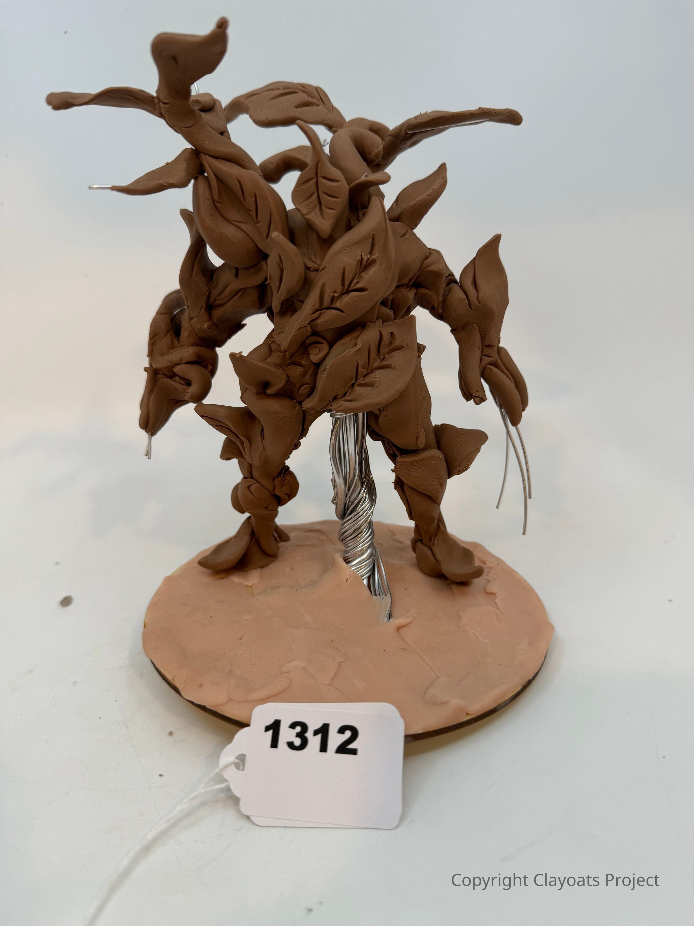 The image depicts a sculpture made of clay shaped like a humanoid figure entwined with leaves and vines, standing on a circular base. The figure has a wire-like structure visible at its core, giving it a sense of form and stability, while the leaves are intricately detailed, showing veins and curvatures that add to the organic appearance. The base is made of a similar clay material and appears slightly textured, suggesting an earthy surface. The overall composition showcases