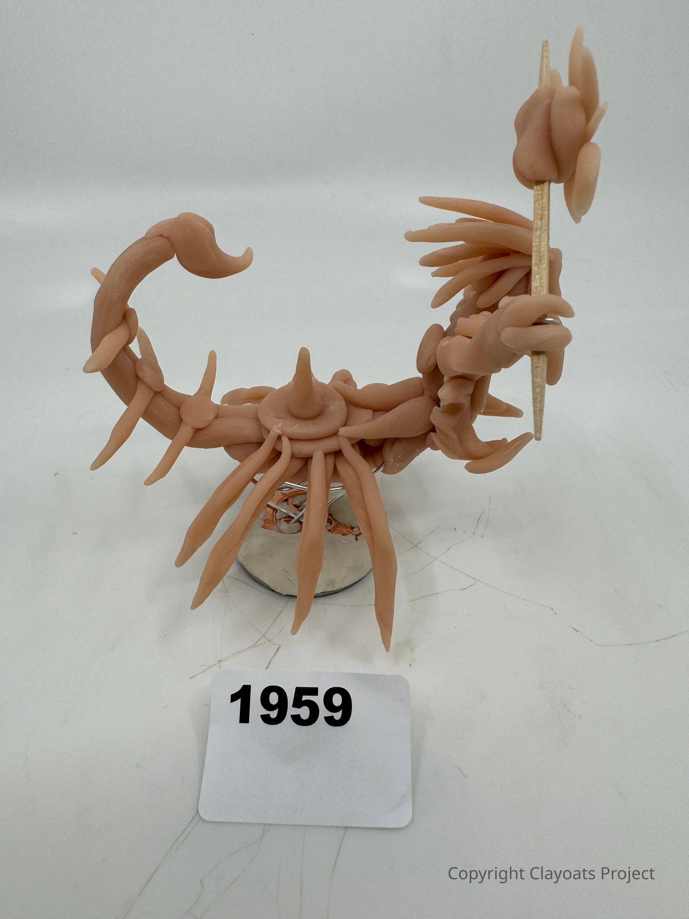 The image shows a creatively crafted sculpture resembling a fantastical creature with numerous elongated limbs and a curled tail, made from a flesh-toned material. The structure appears abstract, with textured details and spikes extending outward, giving it a dynamic, fierce appearance. It holds a staff-like object in one of its limbs, topped with various protruding shapes. The creature is mounted on a cylindrical stand to provide stability, and positioned on a surface with visible scratches.