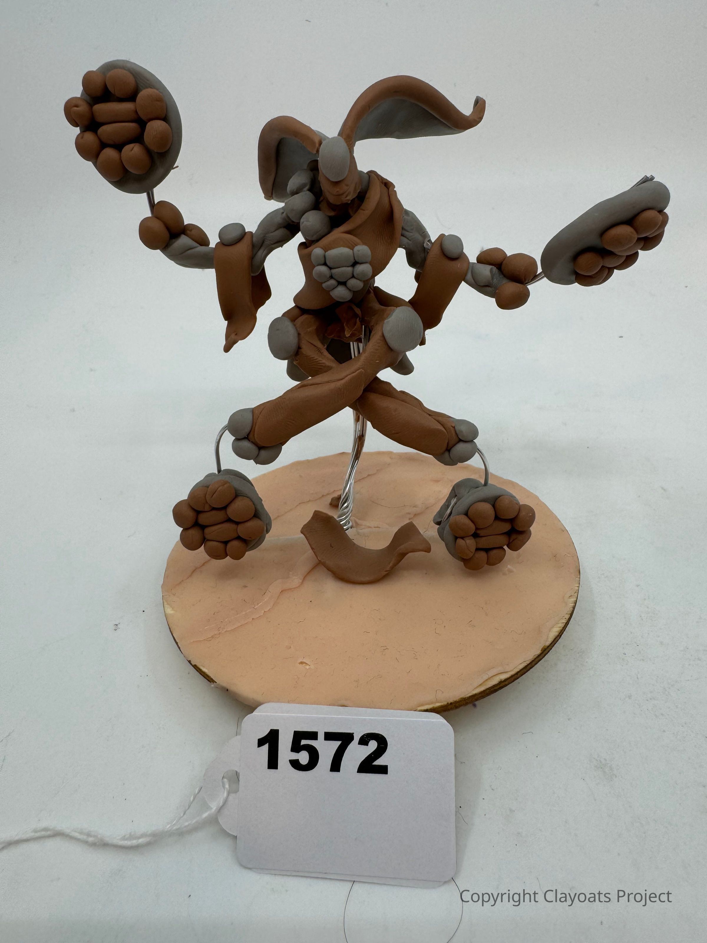 The image depicts a creatively crafted clay sculpture featuring a fantastical humanoid figure with exaggerated proportions and large round fists. The figure is poised in an energetic, dynamic stance, standing on one foot while balancing on a circular base. Its distinct rabbit-like ears and sculpted muscles are highlighted by the contrasting gray and brown tones of the clay. With a sense of motion captured in the image, the artwork suggests a playful character in mid-action, displaying a blend of whimsy and artistry.