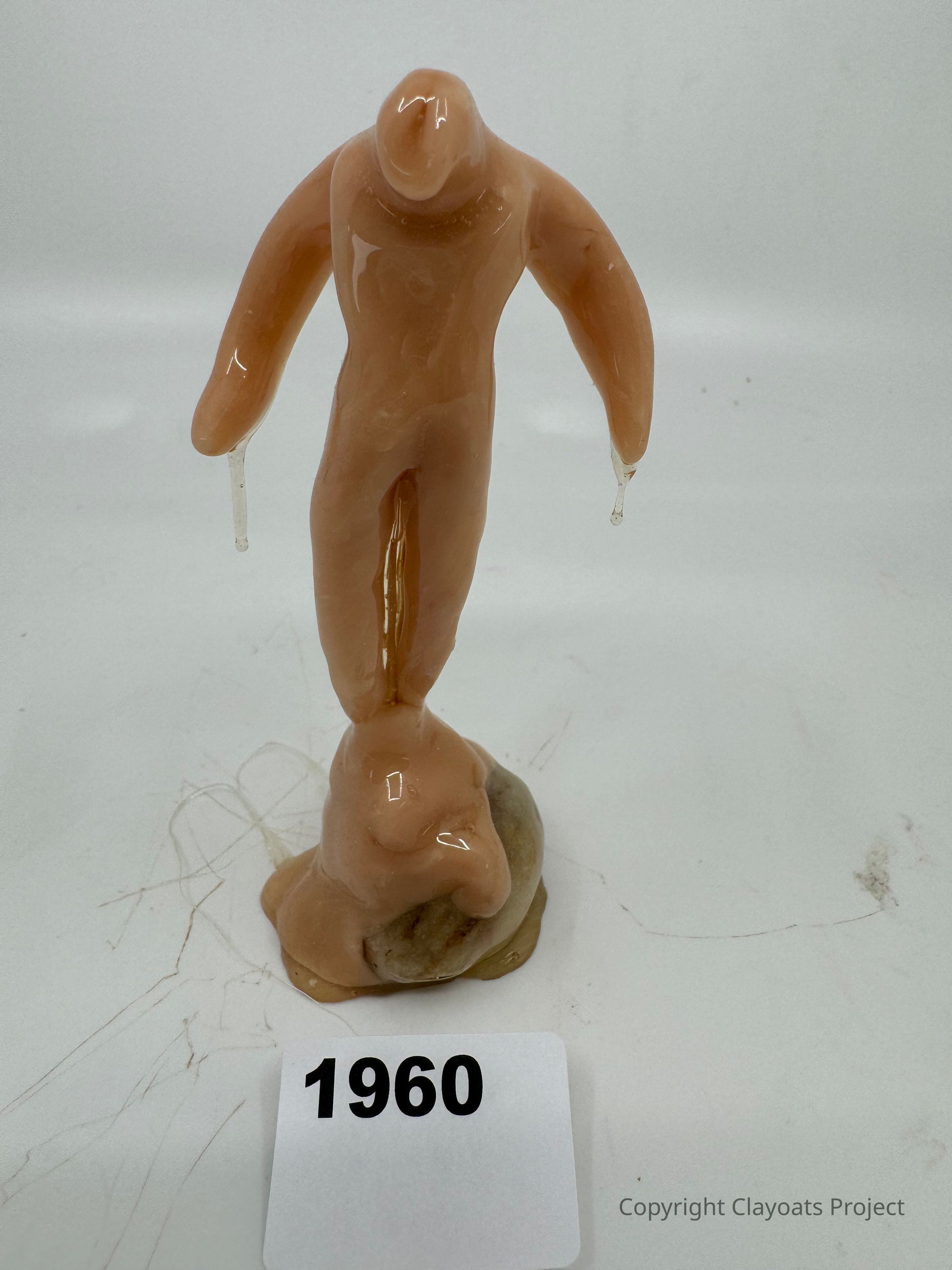 The image shows a wax figure with an abstract human-like form in shades of brown, standing on a lumpy, uneven base. Its arms are outstretched, and it appears to have droplets forming at the ends. The figure's head is featureless, contributing to its minimalist style. The background is plain, placing full emphasis on the wax sculpture and its unique