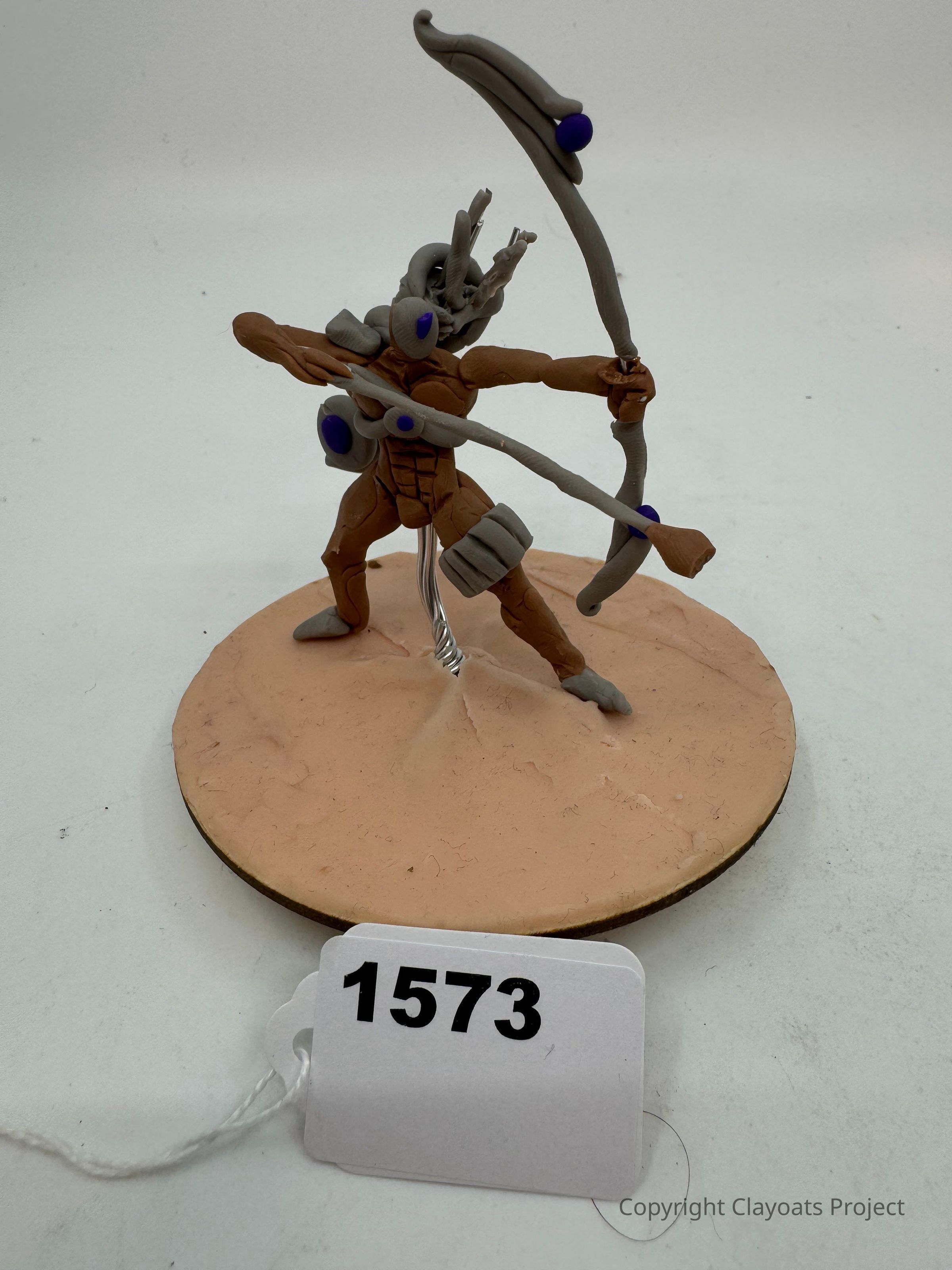 The image displays a handmade, clay figurine of a futuristic archer on a round base, featuring a dynamic pose with a bow and arrow ready to fire, adorned with several purple accents on its armor. The base is light brown. This sculpted figure, with intricate details and a sense of motion, appears to be part of a collection or an art exhibit, capturing the viewer’s attention with its unique design.