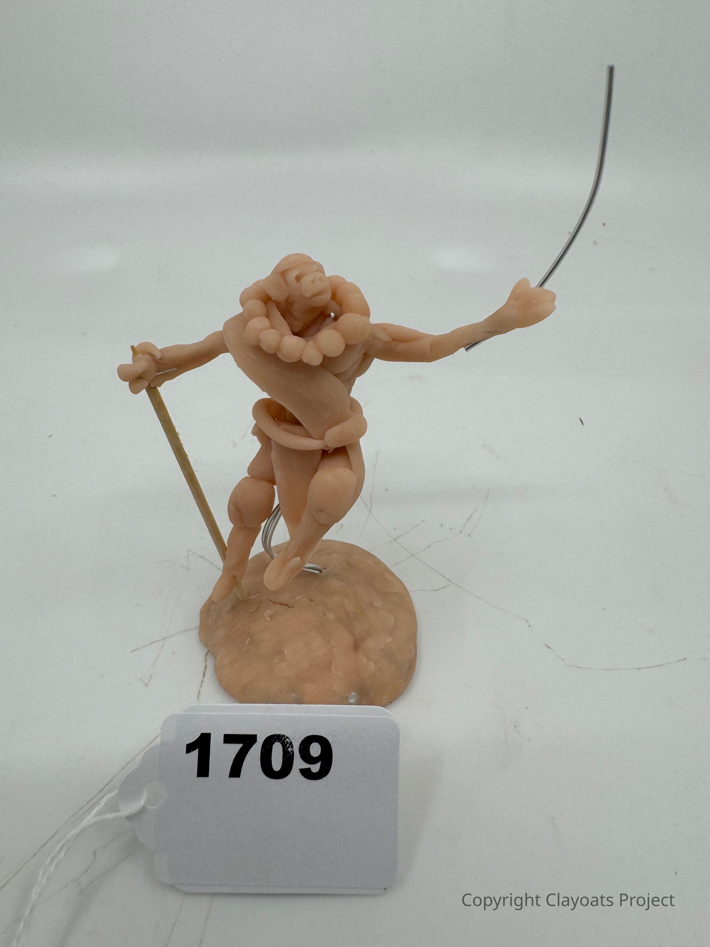 The image depicts a small, clay-like sculpture of a humanoid figure with exaggerated features and a whimsical pose, standing on an uneven base. The figure holds a thin stick in one hand and extends the other arm upward, where it appears to grasp a flexible wire. The sculpture is detailed with intricate, rounded shapes adorning the neck and limbs, adding depth to its form.