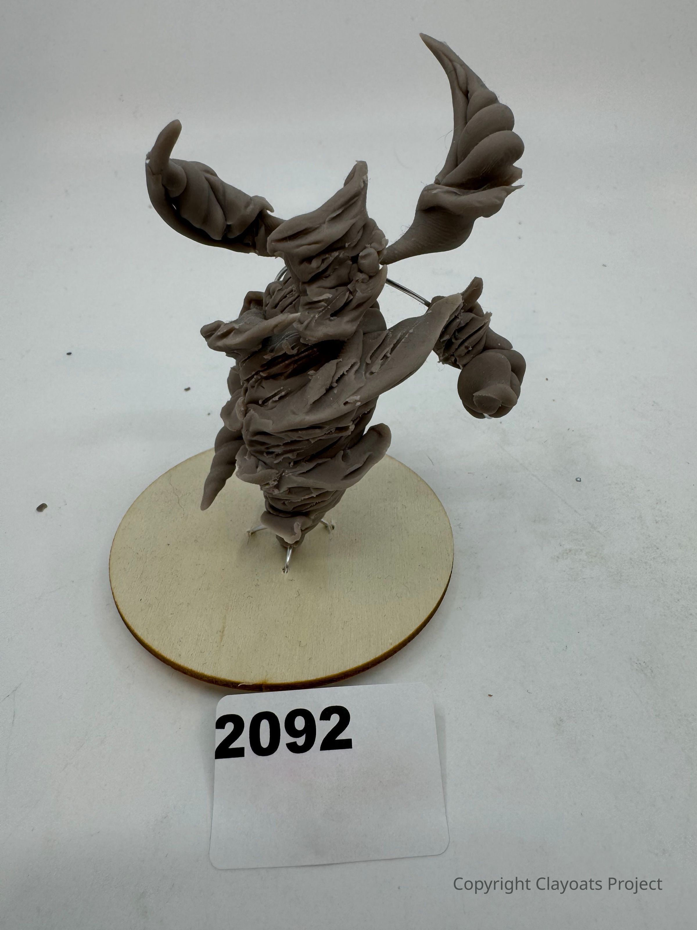 The image displays a uniquely shaped clay sculpture with intricate, twisting forms resembling a fantastical creature or abstract figure, placed on a circular wooden base; nearby, a numbered card with the digits "2092" is positioned, suggesting cataloging or identification. The sculpture's dynamic and textured design creates an impression of movement and energy, hinting at an artistic exploration of organic forms and imaginative expression, while the neutral background emphasizes the artwork’s details, allowing viewers to focus fully on its creativity and craftsmanship.