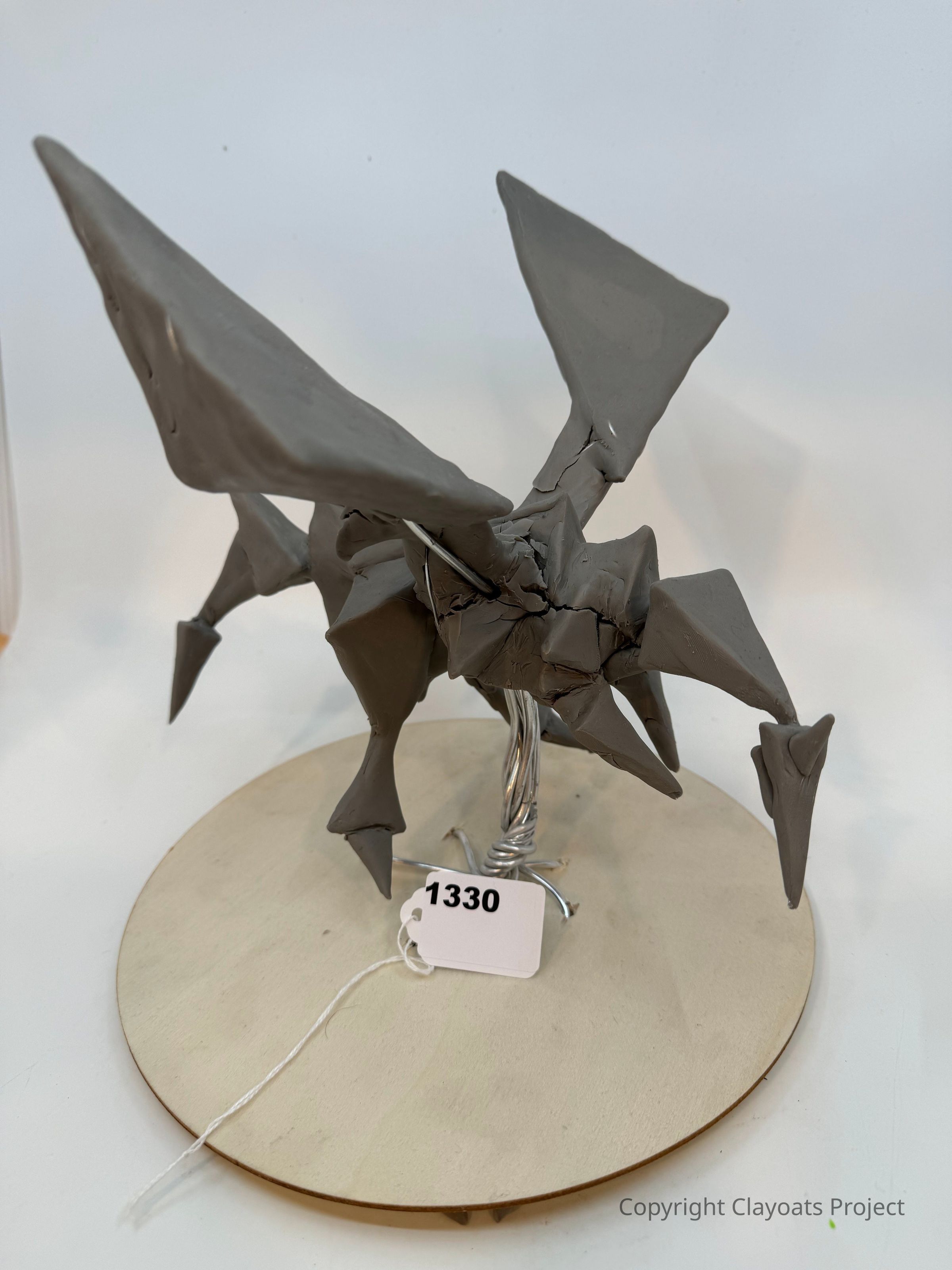 The image depicts a gray clay sculpture of a winged creature with sharp, angular features, resembling a dragon or pterosaur, mounted on a circular wooden base; a metal support helps hold the figure upright, presenting a unique blend of artistic creativity and craftsmanship, all set against a plain white background that highlights its intricate details and dynamic form, capturing the viewer's eye.