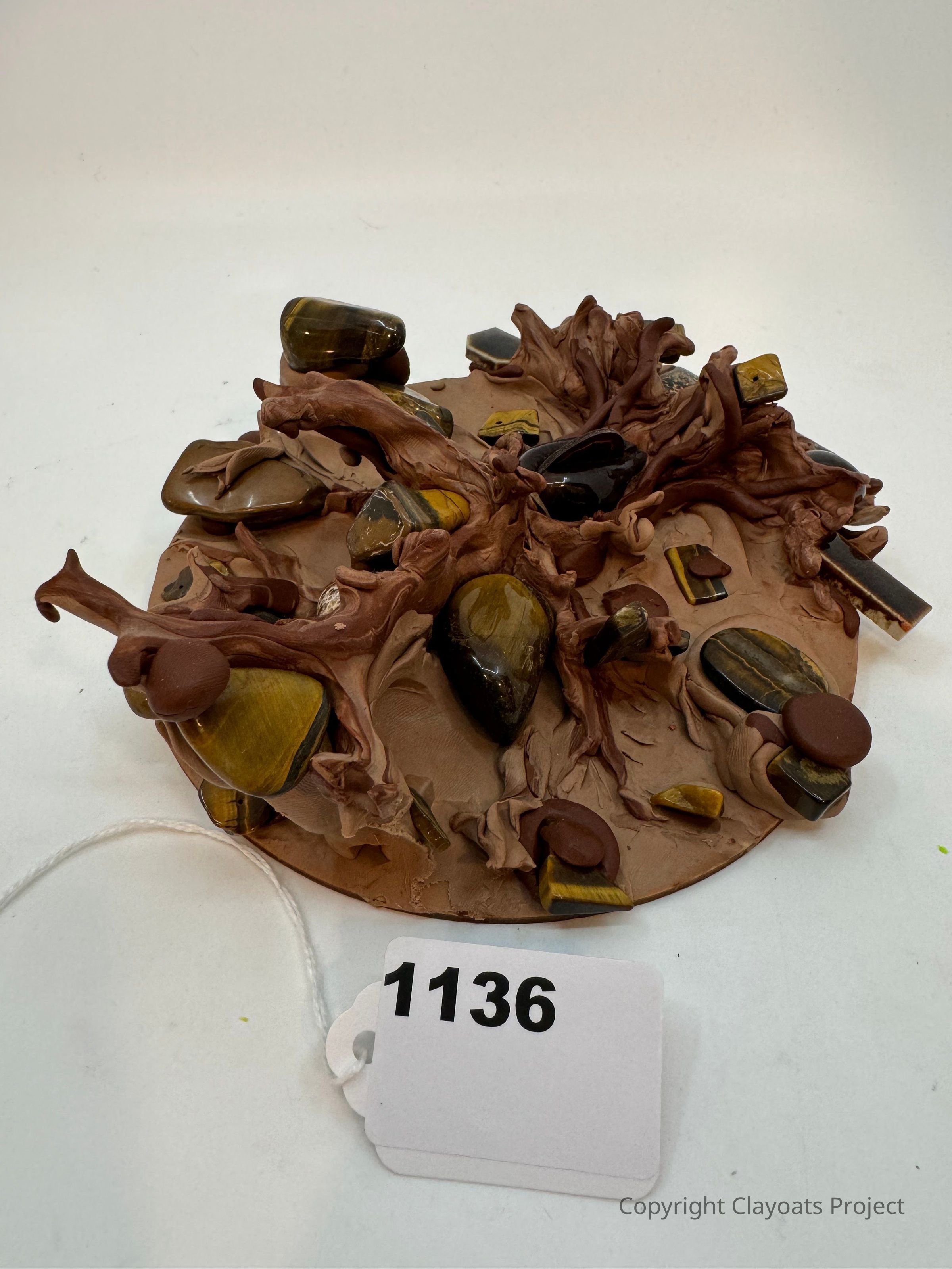 The image displays a round clay sculpture on a white surface, featuring intricate, branch-like formations adorned with polished, multi-hued stones embedded throughout, creating a naturalistic, organic appearance. The combination of earthy tones and smooth textures gives the piece a tactile, visually interesting quality, suggesting themes of nature and craftsmanship intertwined harmoniously through artistic expression and meticulous design.