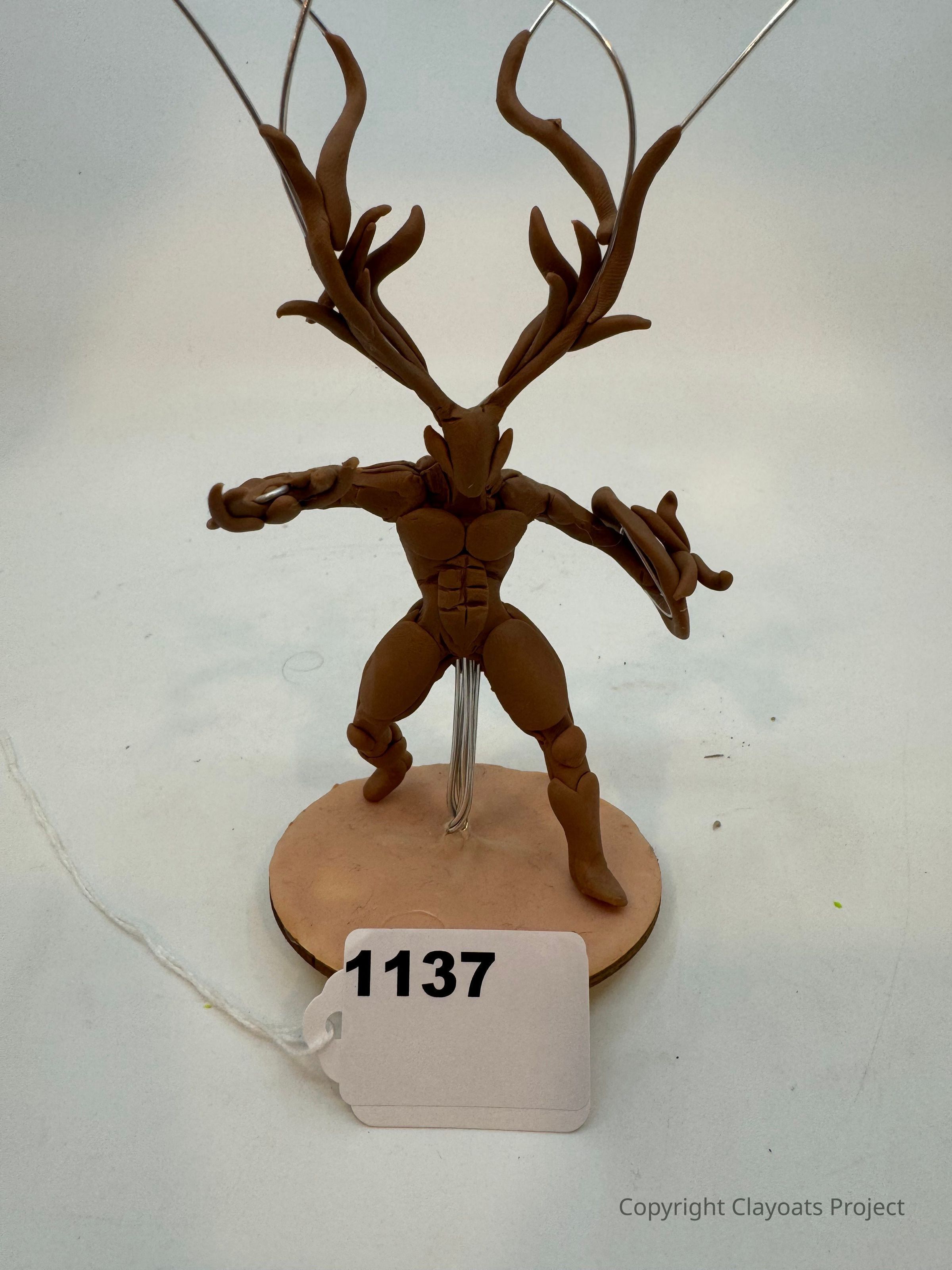 The image shows a handcrafted sculpture made from brown clay depicting a humanoid figure with elongated, tree-like antlers that extend upwards and outwards. The figure stands in a dynamic pose on a circular base, with a wire visibleq supporting the structure from behind. The sculpture's intricate details and stylized antlers give it an imaginative and artistic appearance, potentially representing a mythological or fantastical creature.