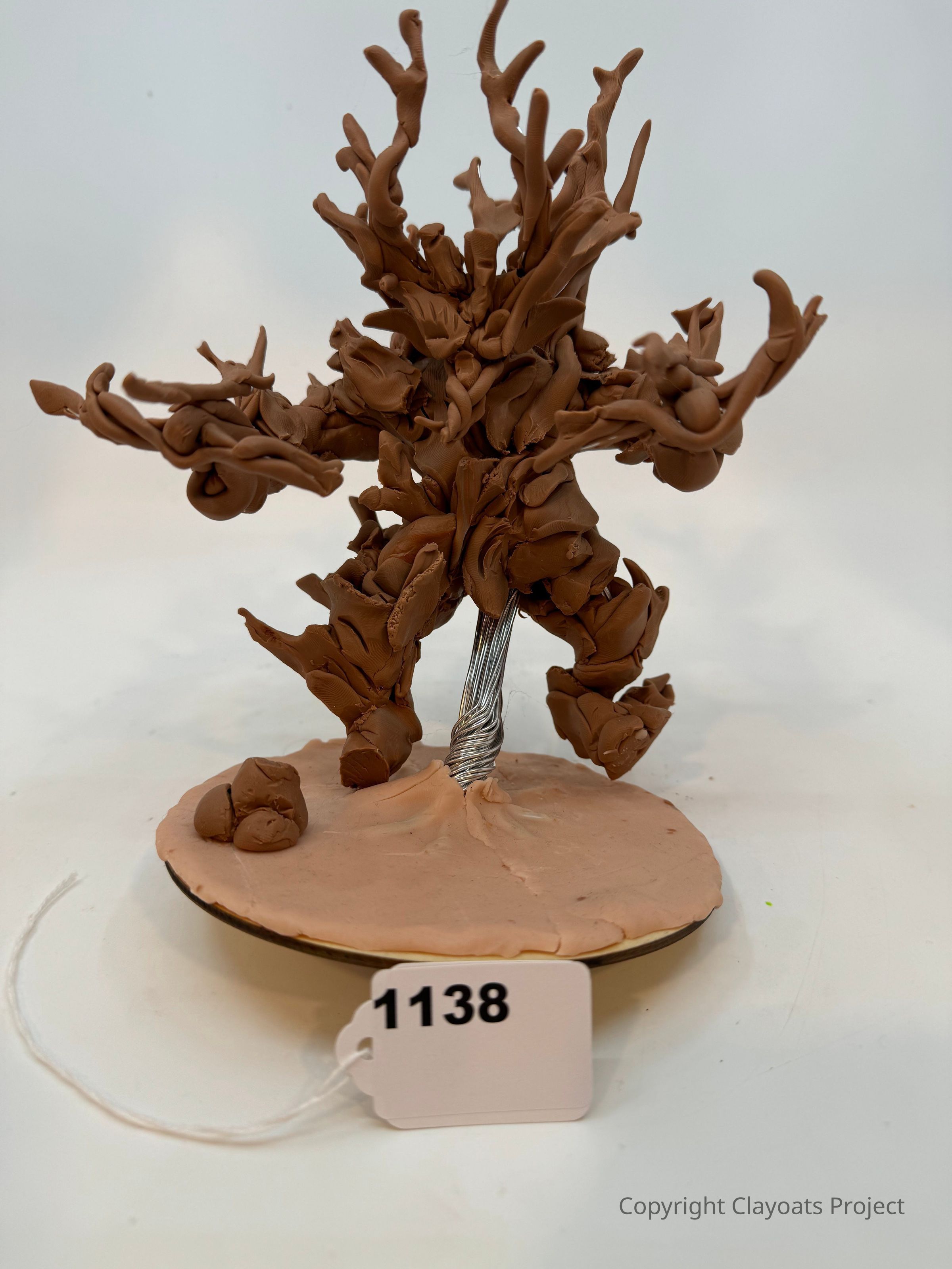 The image depicts a creative sculpture made from brown clay, resembling an abstract or fantastical figure. It stands on a round base, with a metal wire visible at its center. The sculpture is characterized by its intricate, swirling shapes and limb-like extensions, giving it an organic appearance. It occupies a neutral background, suggesting focus on the artwork itself.
