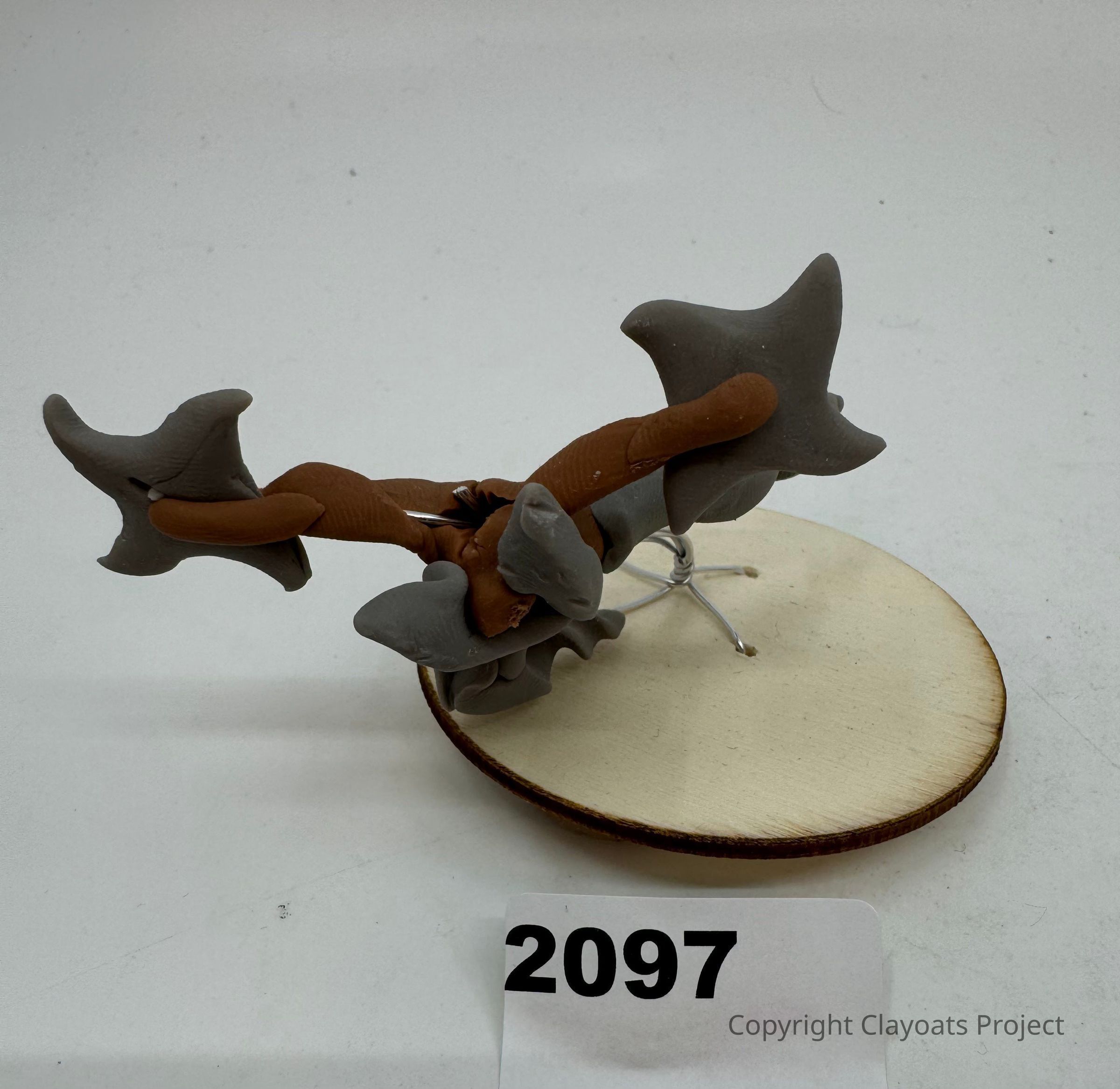 The image features a unique, abstract sculpture composed of interwoven brown and gray elements, resembling organic shapes or creatures, placed on a circular wooden base with a metal wire support, all set against a plain background; a small label with the number "2097" is positioned in front, possibly indicating cataloging or reference information.