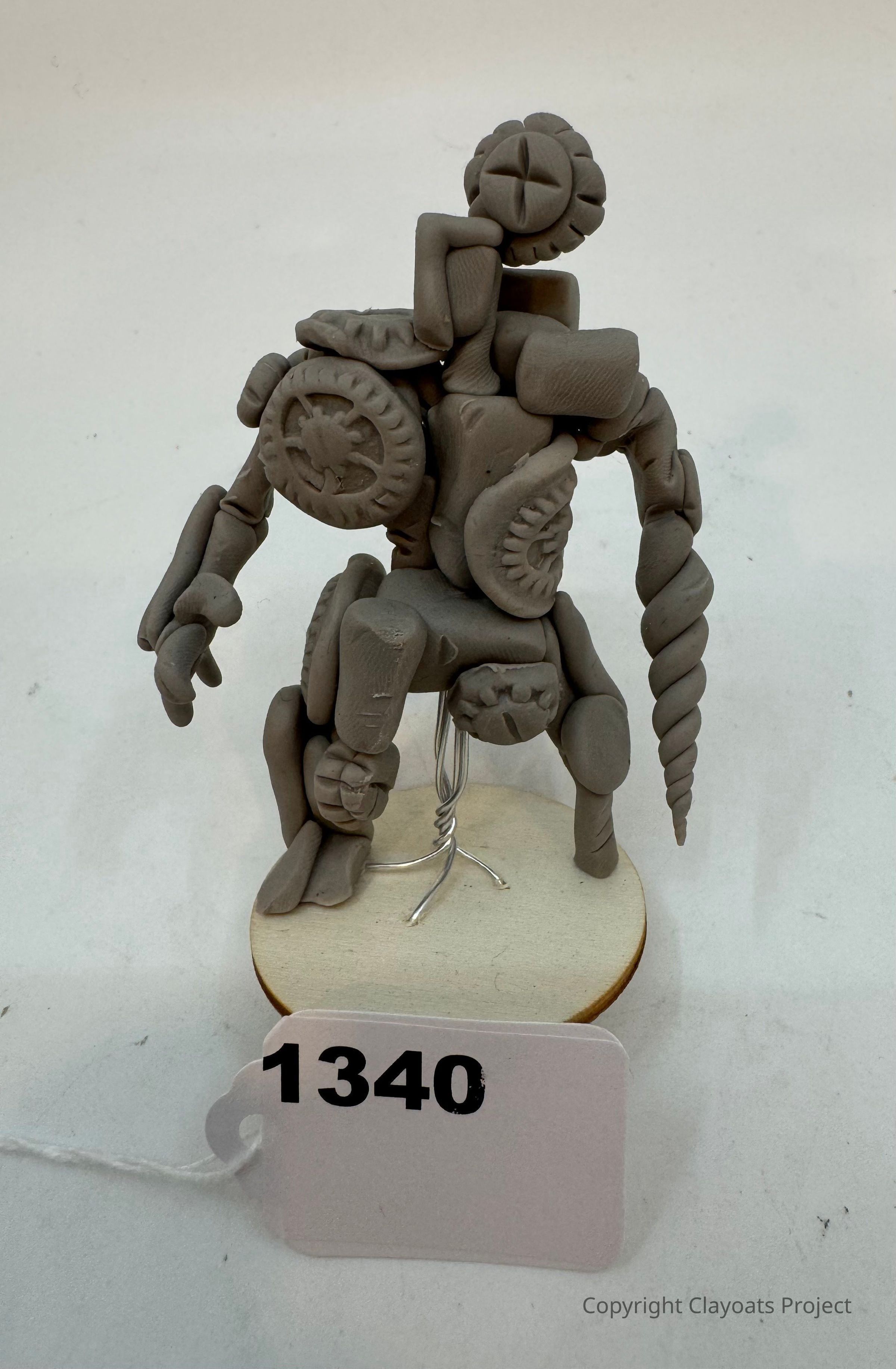 The image depicts a uniquely crafted sculpture made from a grey, clay-like material, featuring various geometric shapes and intricate designs, resembling a humanoid figure displayed on a wooden base; its components include circular and cylindrical forms, with some surfaces displaying star-like patterns, while a spiral element extends from the right side, creating an impression of a fantastical creature; the sculpture is presented on a stand, stabilized by a visible wire.