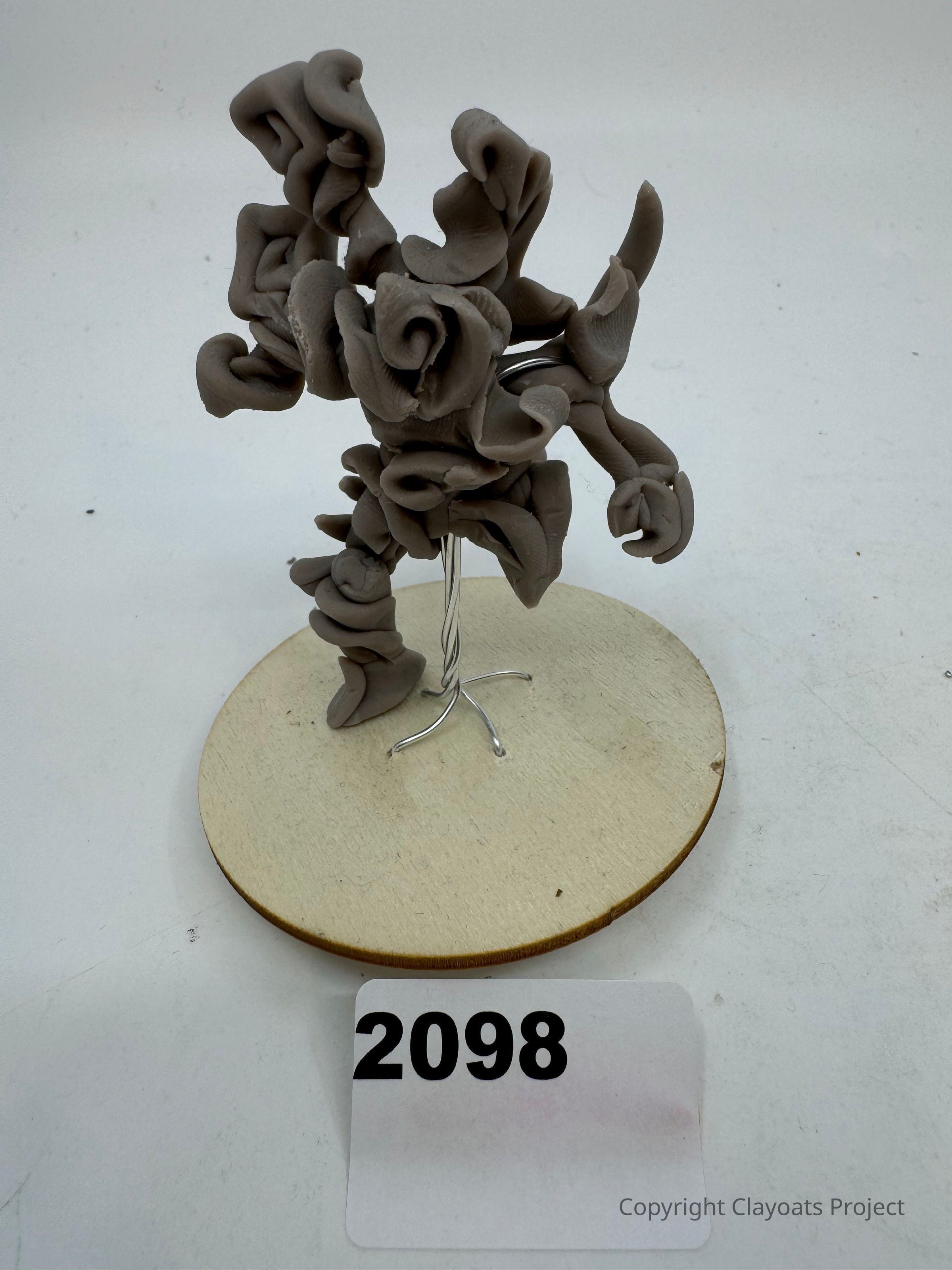 The image showcases a sculptural piece made from twisted and coiled clay, artistically arranged on a round wooden base, held together by a metal wire frame. Placed on the surface in front of the sculpture is a card displaying the number "2098" in bold black text. The entire composition sits against a plain white background, highlighting the intricate details and organic shapes of the clay, which evoke a sense of fluid motion and abstract form.