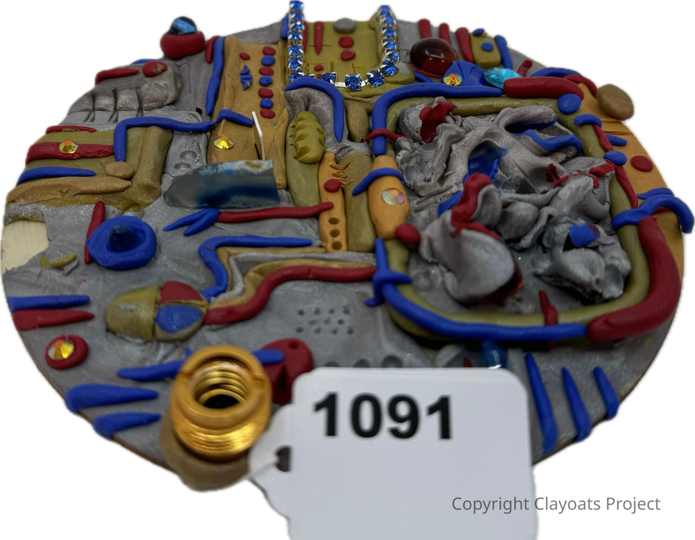 The image showcases a round, abstract sculpture made from various materials, featuring intricate patterns and designs in colors including red, blue, gray, and gold, adorned with decorative elements like shiny rhinestones and textured sections, some of which resemble pipes and organic shapes; prominently displayed in the lower center is a white tag labeled "1091," adding an element of identification to the otherwise whimsical and artistic piece.