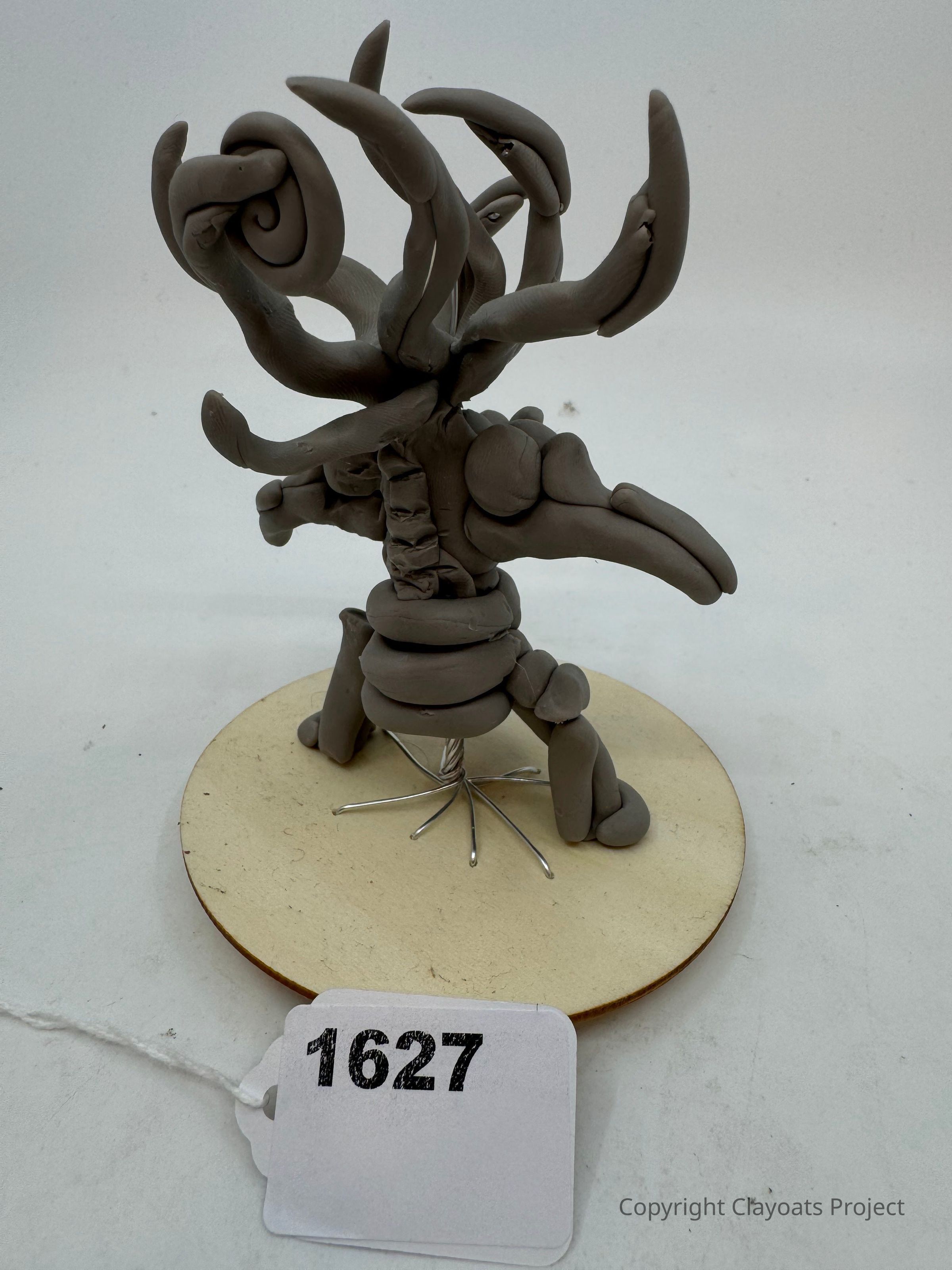 The image depicts a gray clay sculpture with intricate, swirling designs that resemble tentacle-like structures extending outward from the center, which is mounted on a circular base. The sculpture stands on what appears to be a wire coil, The abstract and artistic nature of the creation evokes a sense of creative imagination, while the craftsmanship highlights the artist’s skill in manipulating the clay into this unique form.