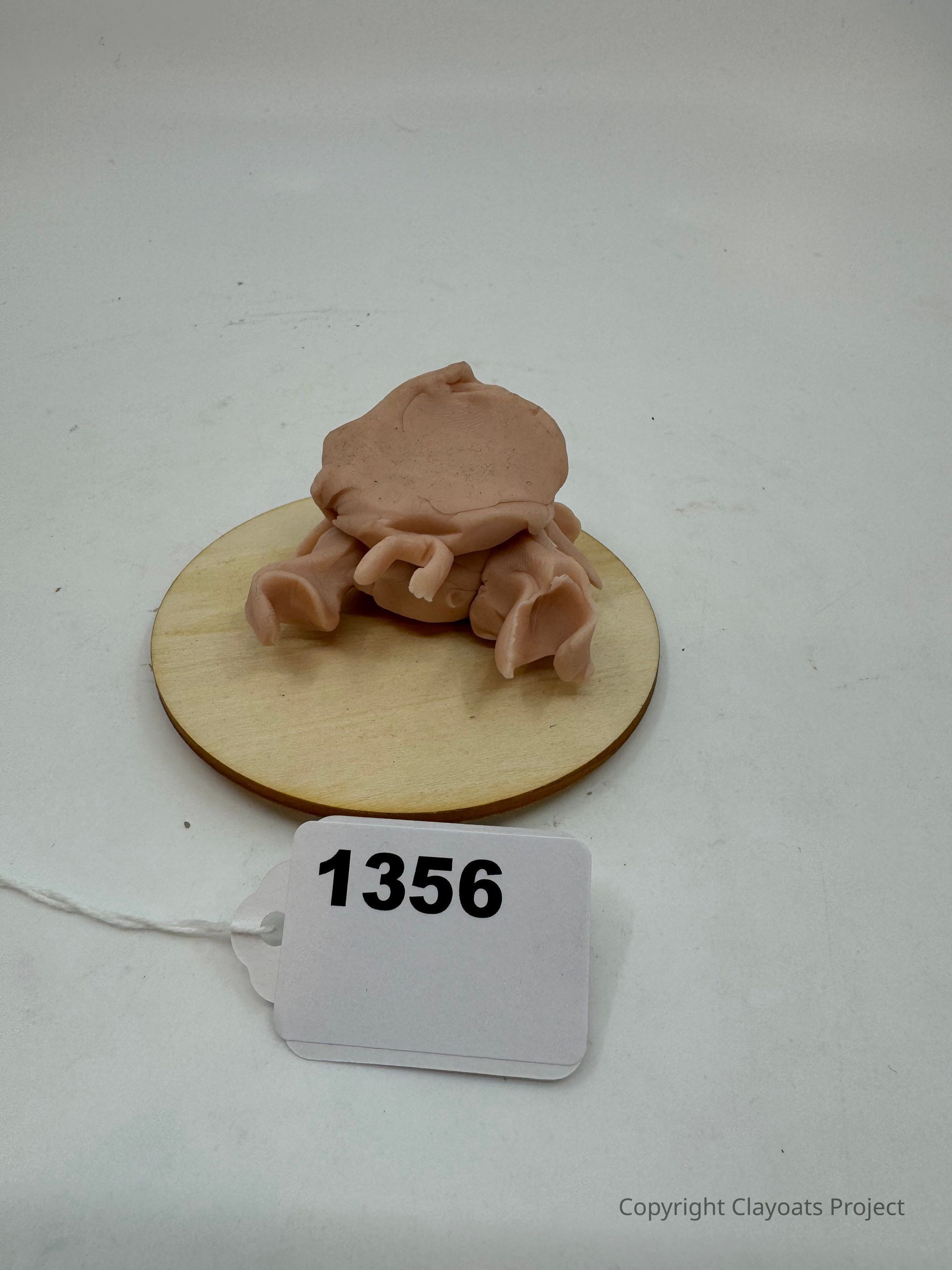The image shows a small, abstract sculpture made of a soft, clay-like material, resting on a round wooden base. The sculpture appears to be organically shaped, possibly mimicking a creature or object, emphasizing its handcrafted and unique nature, highlighted by the neutral background that ensures the focus remains solely on the object and its details. The entire setting provides a minimalistic and