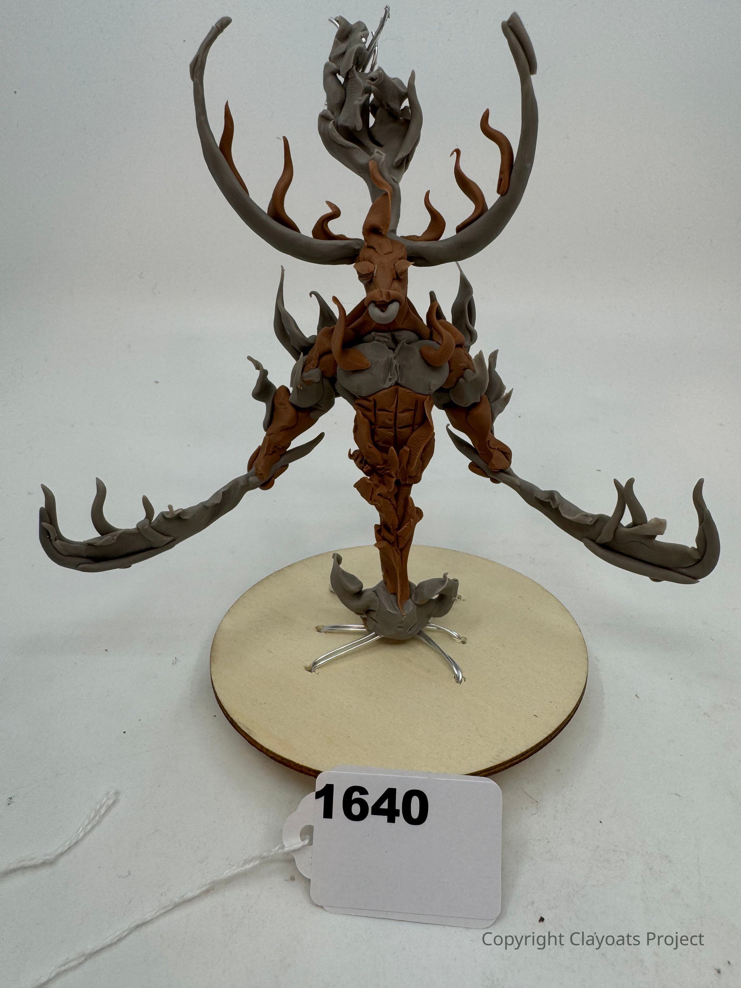 The image depicts a unique sculpture of a fantastical creature made from materials like clay, featuring elongated limbs, antler-like structures, and intricate details resembling swirling flames or vines. This imaginative figure is positioned on a circular base with metallic supports underneath, giving it a dynamic and mystical appearance.