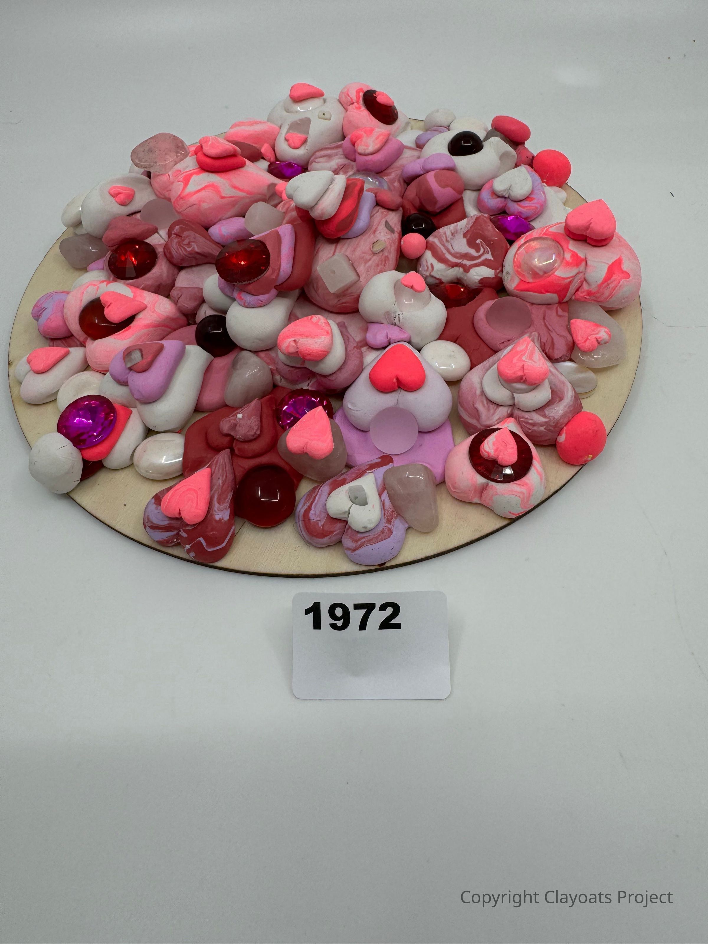 The image features a circular arrangement of various pink, red, and white heart-shaped and abstract clay or stone objects on a round wooden base. The objects are interspersed with vibrant, shiny gemstones in red and purple hues, creating a playful and vibrant aesthetic. The assortment includes swirled patterns and smooth, glossy textures, enhancing the visual interest.