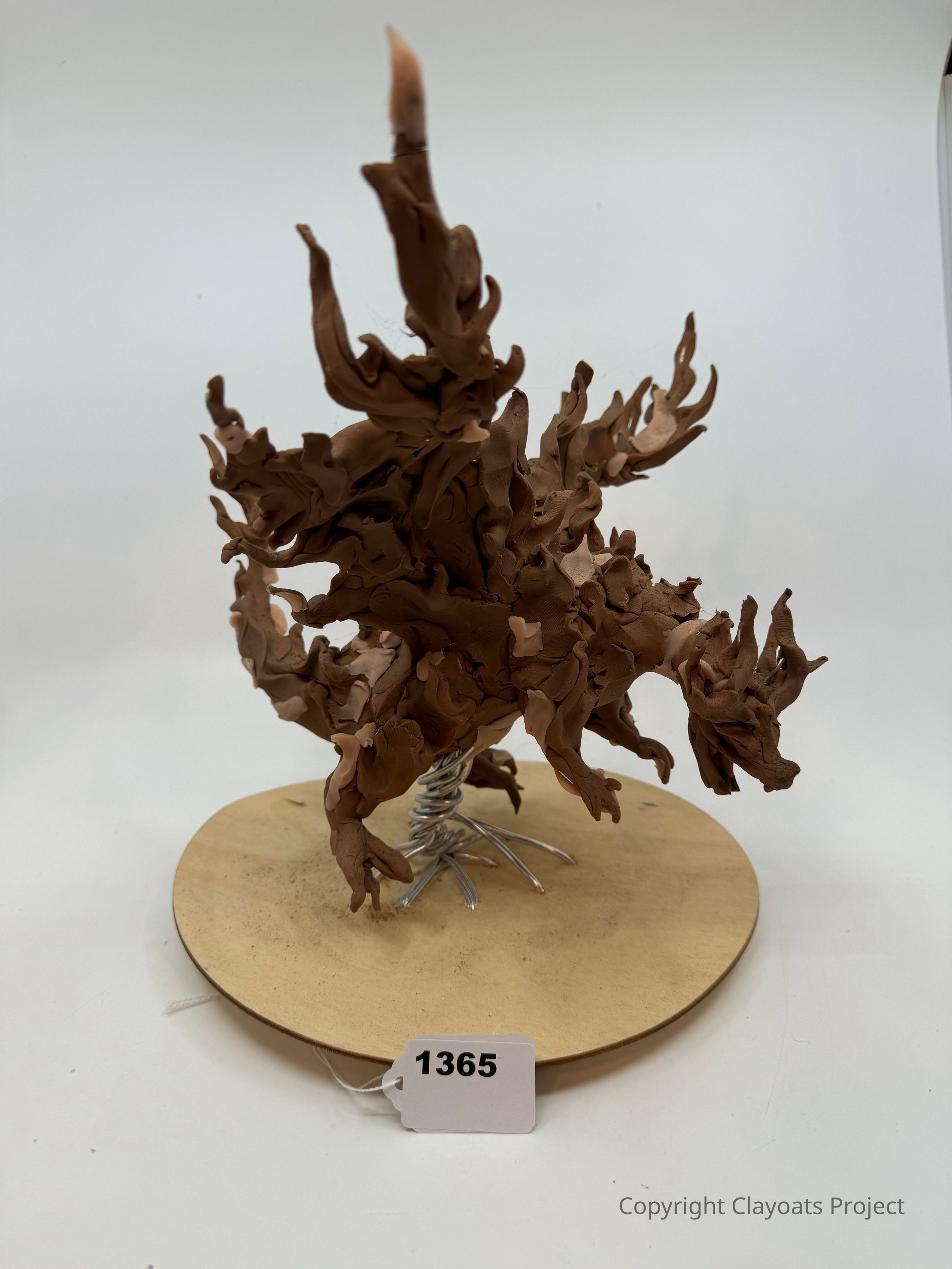 The image shows a dynamic, abstract clay sculpture resembling a swirling, flame-like formation with intricate and textured detailing. It is mounted on a round wooden base supported by a coiled metal structure, suggesting movement and vitality. The sculpture is predominantly earthy brown.. The background is a plain, neutral white, allowing the sculpture’s complex shapes and curves to stand out prominently without