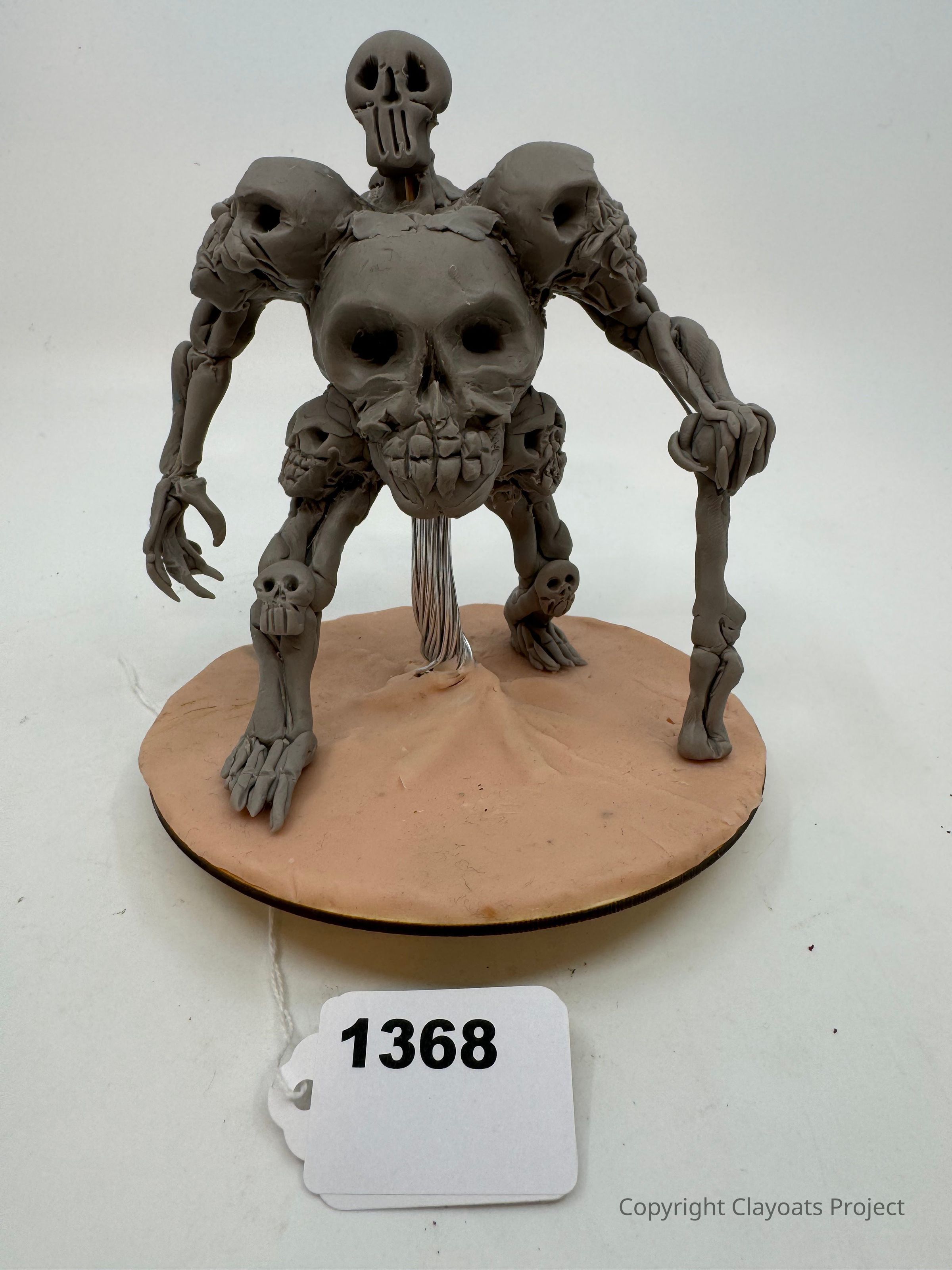 The image features a clay sculpture of a multi-headed skeletal creature with skulls integrated into its form, standing on a circular base resembling sand or earth. The sculpture is detailed, with elongated limbs and emphasized skeletal features, giving it a menacing appearance. Its primary skull is central and prominent, with hollow eye sockets, while additional skulls protrude from the shoulders and joints, creating a haunting, otherworldly presence.